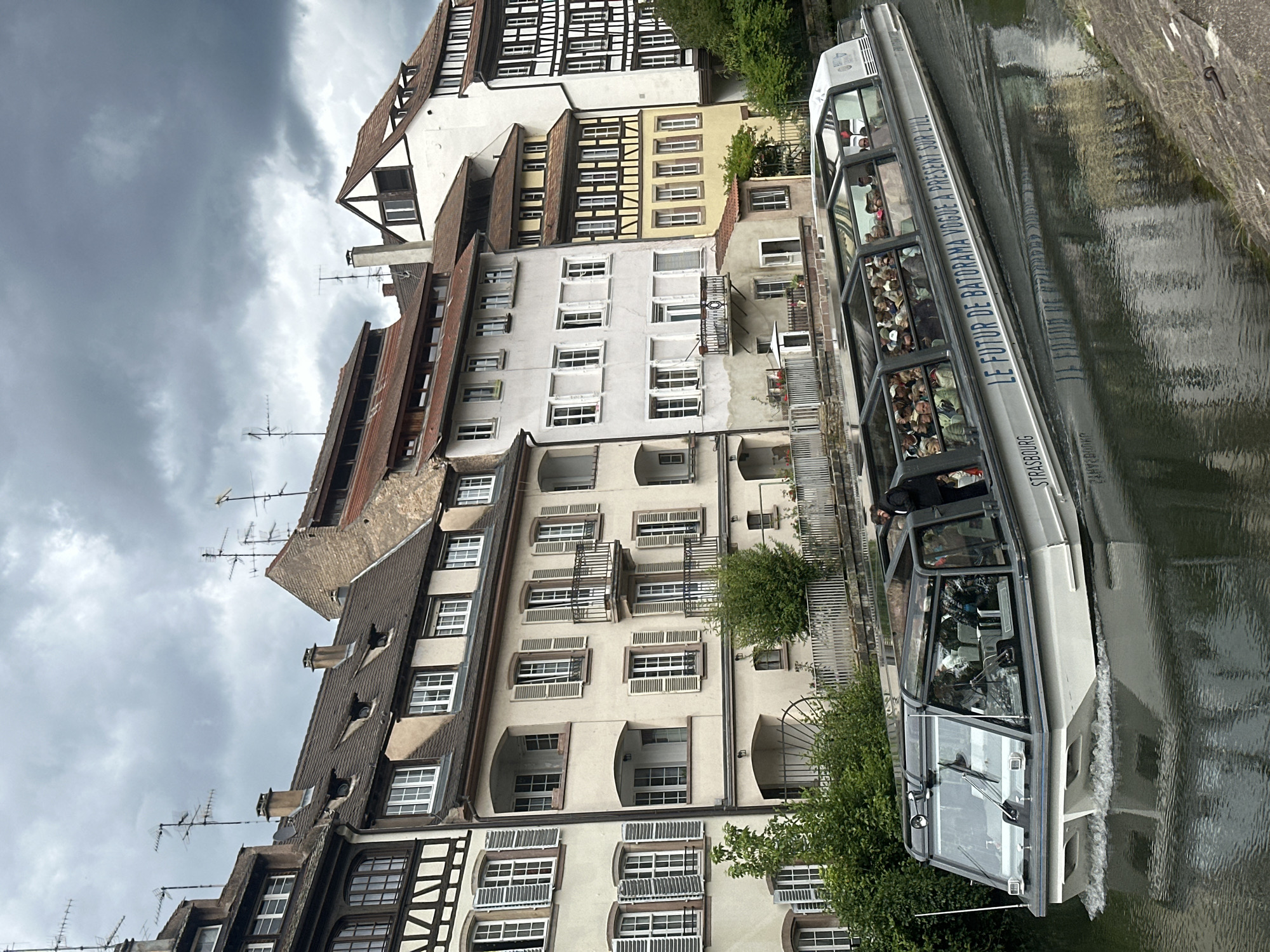This image depicts a tour boat navigating a canal in Strasbourg, France. The boat, which is silver and has a glass roof, is filled with passengers and is branded with the text "LE FUTUR DE BATODRAMA" and "STRASBOURG" on its side. The buildings lining the canal are characteristic of Strasbourg's historic architecture, featuring half-timbered facades, various window styles, and numerous chimneys. The sky is overcast, suggesting a cloudy day. The water in the canal reflects the surrounding buildings and the boat.