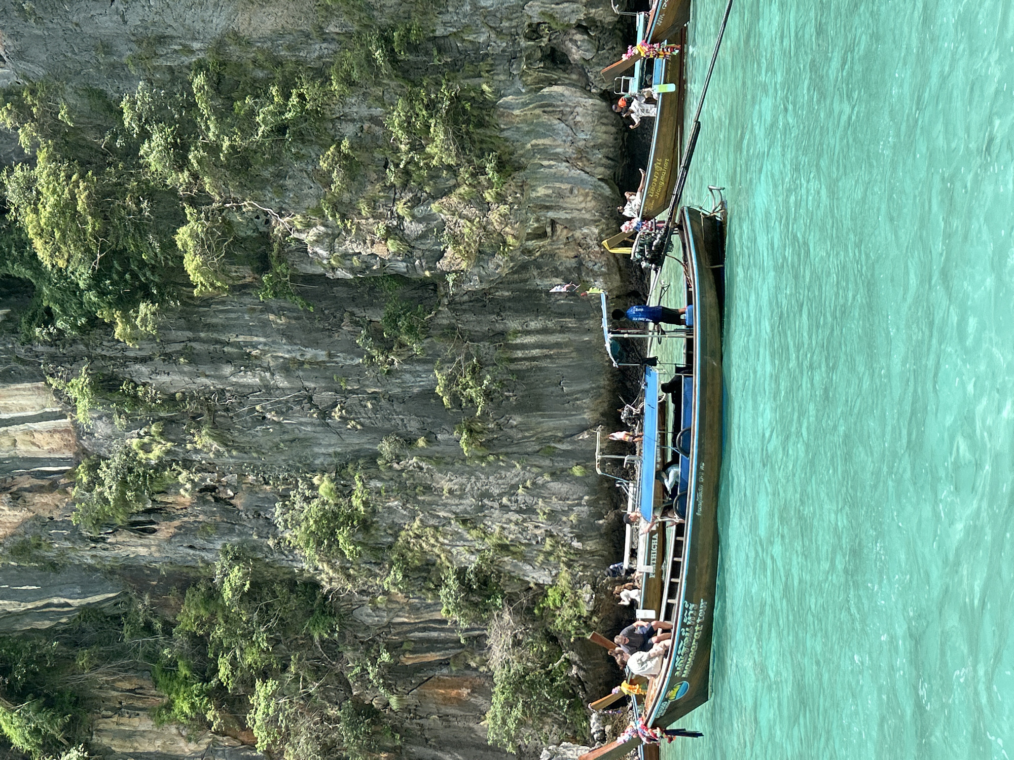 A vertical, eye-level, medium shot captures a vibrant scene of turquoise water and a sheer limestone cliff face covered in dense green foliage. Several traditional long-tail boats, characteristic of Ao Nang, Thailand, are anchored in the water. The foremost boat, brown with a blue stripe, has passengers seated on its edge. The water is a vivid, clear turquoise, with small waves and ripples on its surface. Behind this boat, another long-tail boat, with a blue canopy, is visible, also with passengers. Further back, more boats are present, some partially obscured by the cliff. The cliff itself is imposing and textured, with numerous small trees and plants growing from its crevices. The overall impression is one of a scenic, tropical location with typical Thai tourist activity.
