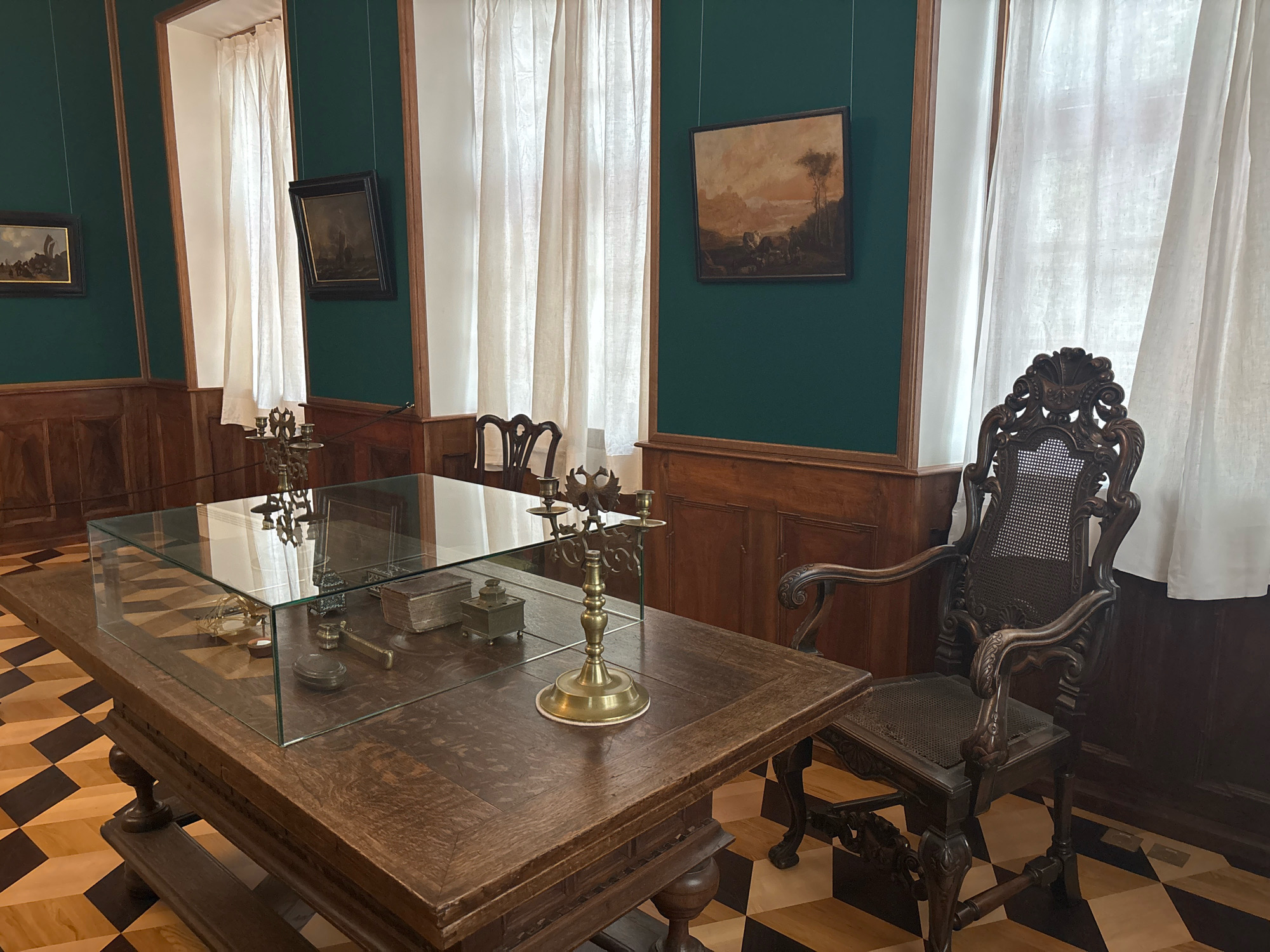 The image depicts a display of historical artifacts within a room that appears to be a museum exhibit, possibly located in Saint Petersburg, Russia. The room features dark green walls and wooden paneling. Several framed paintings are hung on the walls, depicting landscapes and maritime scenes.
In the foreground, a large, dark wooden table with ornate carvings holds a glass display case containing various objects, including what appear to be antique writing instruments, small boxes, and a candelabra. A second, identical candelabra stands on the table outside the display case. To the right of the table is an elaborately carved wooden armchair with a cane seat.
The floor is a distinctive checkerboard pattern of light and dark wood. Large white curtains hang from tall windows on the right side of the room, partially obscuring the view outside. The lighting suggests an indoor setting, possibly with natural light filtering through the curtains. There are no people visible in the image.