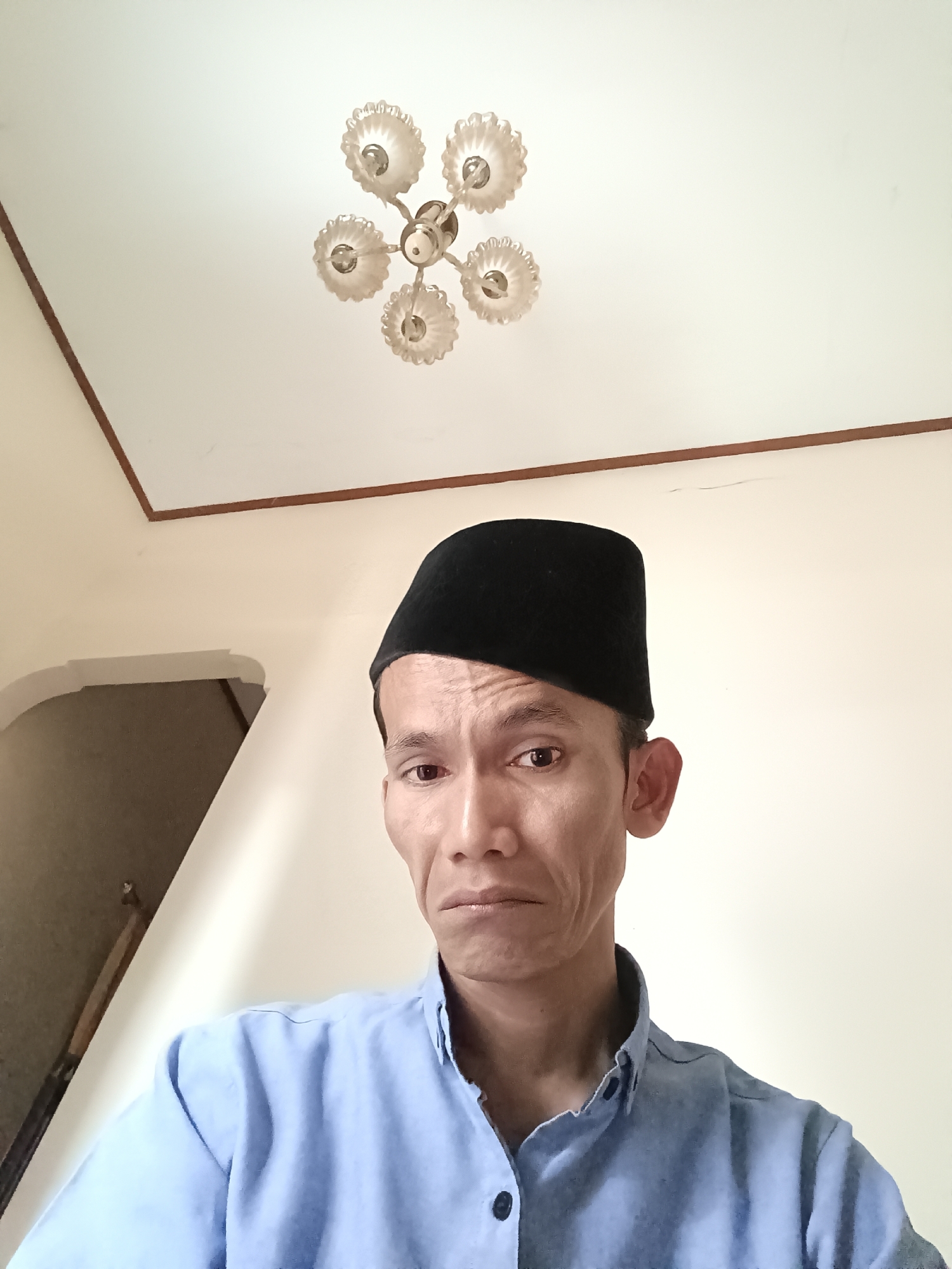A man wearing a black peci and a light blue collared shirt is photographed from the chest up, facing the camera directly. He has a serious expression. In the background, a white ceiling with a decorative light fixture is visible. To the left, a portion of a wall with an arched opening is also present. The overall environment appears to be indoors. There is no specific indication of the location being in Kediri City, Indonesia.