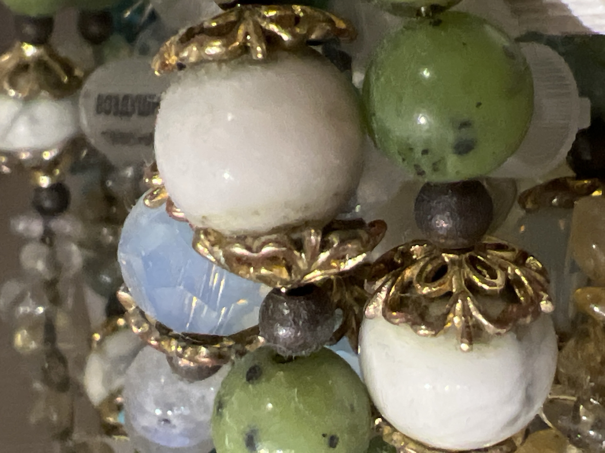 The image displays a close-up, macro shot of a beaded bracelet. The bracelet is composed of various beads, including round white beads, green speckled beads, and a faceted light blue bead. There are also dark, metallic-looking beads and ornate gold-colored bead caps. The background is blurred, showing more beads and possibly other jewelry. There are no people or any recognizable elements of the environment visible, making it impossible to determine a location in Sinyavino, Russia, or any other geographical context.