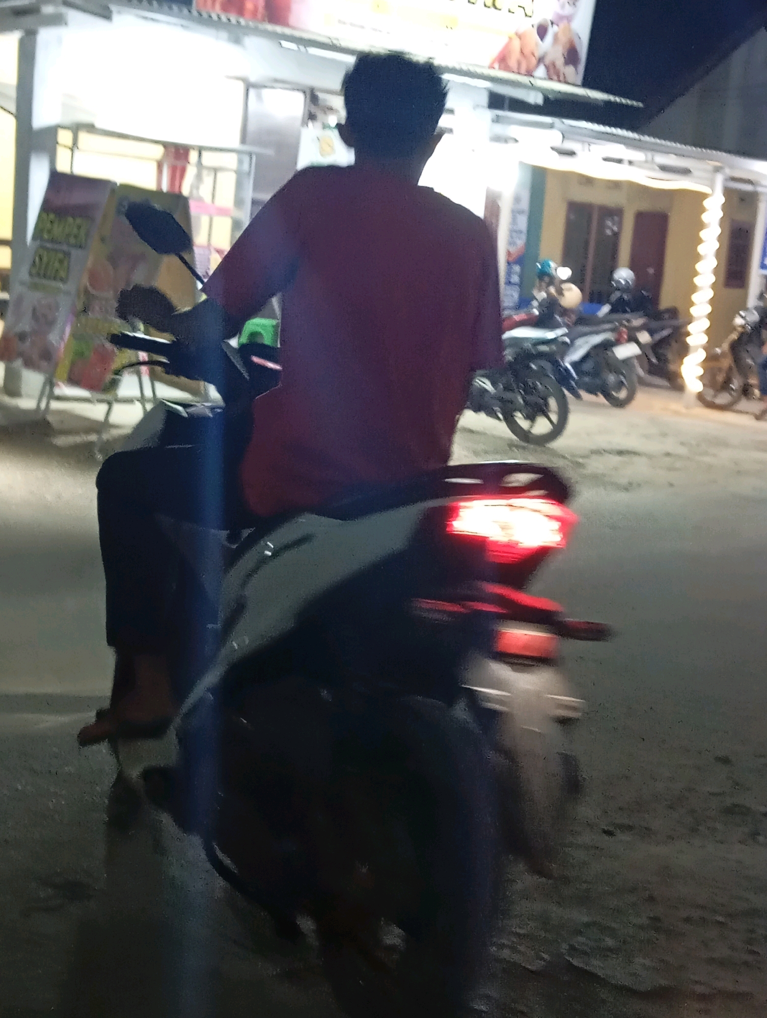 The image captures a nighttime scene on a street in Leman Benawi, Indonesia. A person wearing a red shirt is riding a motorcycle. The motorcycle has a lit red taillight, indicating it is in motion. In the background, several other motorcycles are parked or also in motion. Storefronts with illuminated signs are visible, suggesting a commercial area. The street appears wet, possibly from recent rain, with reflections visible on the surface. The overall lighting is dim, with artificial lights from the storefronts and the motorcycle's taillight providing illumination.