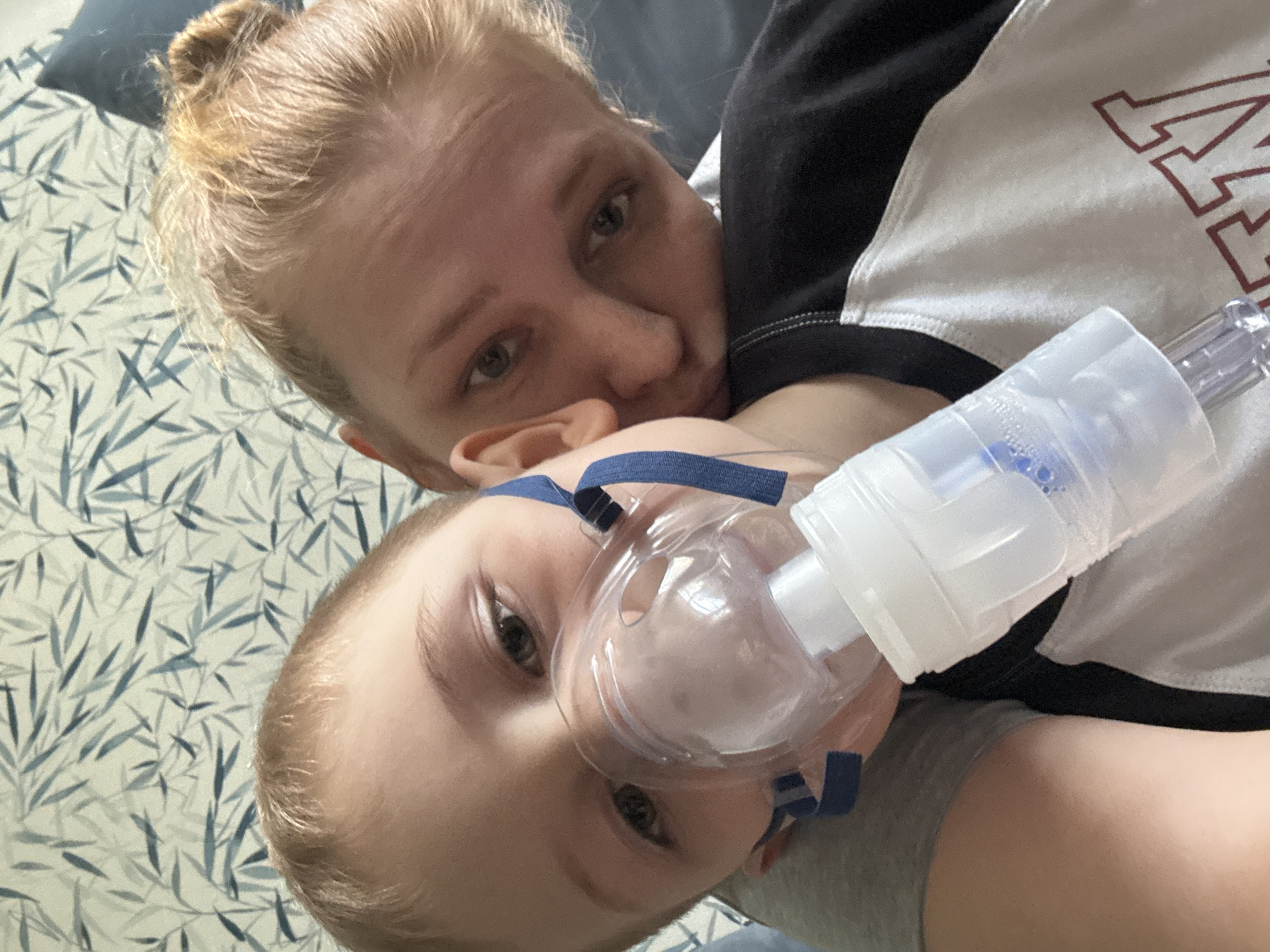A woman with blonde hair in a bun is lying on a bed with a young child. The child is wearing a nebulizer mask. The woman is wearing a white shirt with black sleeves and a red letter "M" on the front. The bed has a blue and white patterned bedspread. The location is not provided.