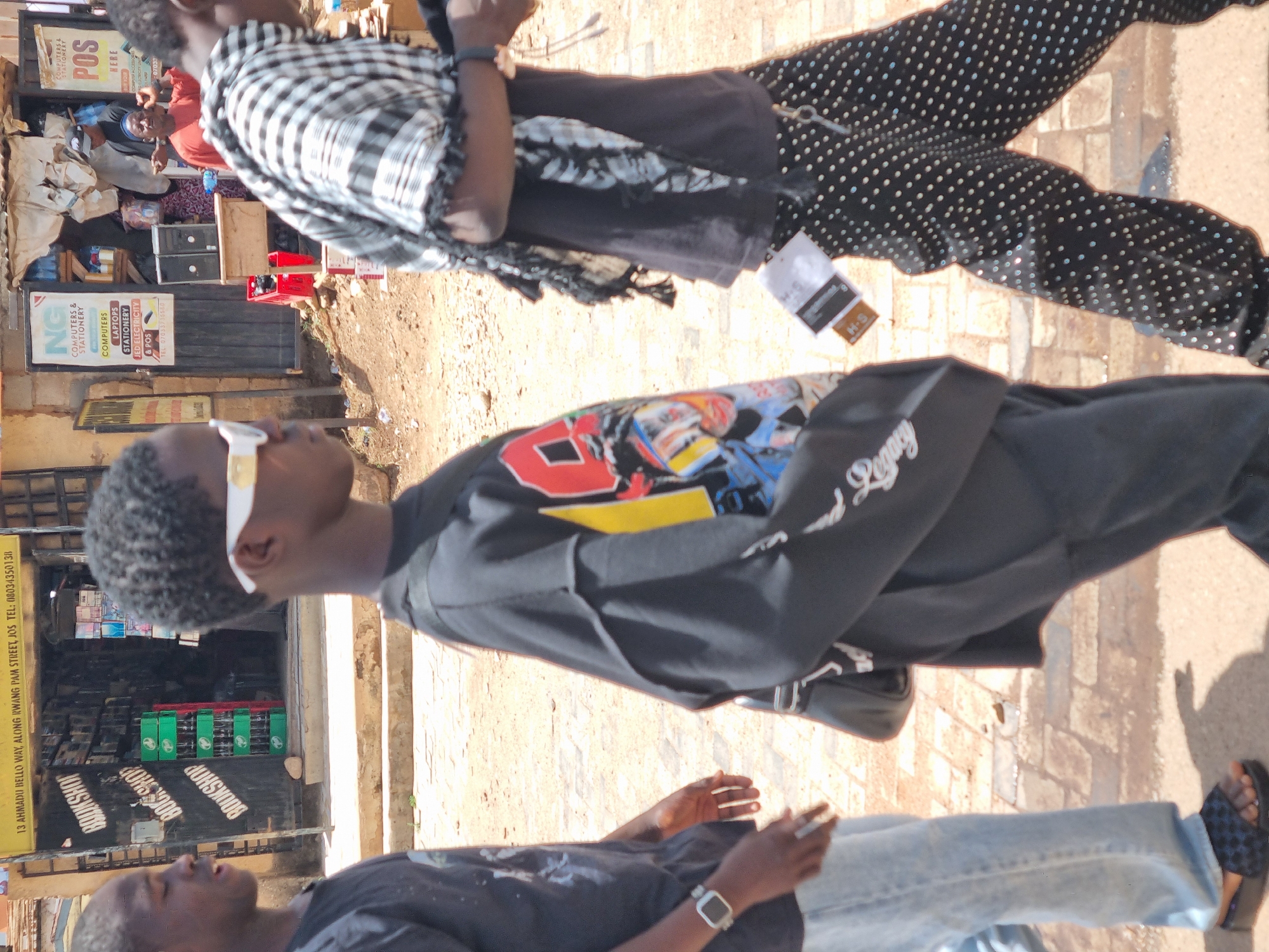 The image shows a street scene in Jos, Nigeria. The location is identified by the sign "13 Ahmadu Bello Way, Along Irwang Pam Street, Jos Tel: 08034350138." The street appears to be paved and has a shop called "Bookshop" with multiple green boxes arranged in front of it. Adjacent to the "Bookshop" is "NG Computers & Stationery." Several individuals are present in the scene. One person is wearing sunglasses, a black shirt with a colorful design and the word "Legacy" on the sleeve, and black shoes. Another individual is wearing a black and white checkered garment and black pants with white polka dots. A third person is wearing a dark t-shirt, a watch, and light blue jeans.