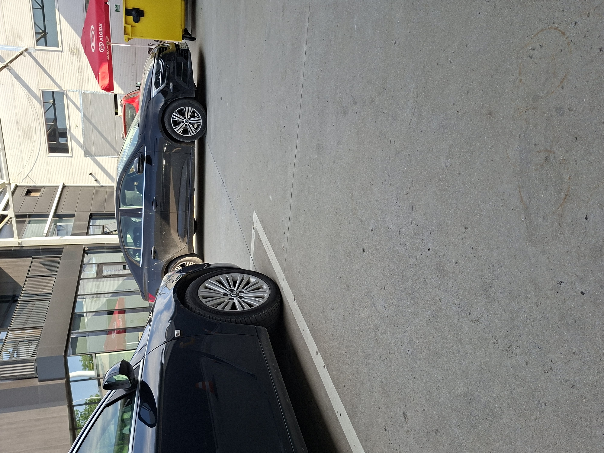 The image shows two dark-colored cars parked in a parking lot. The cars appear to be adjacent to a modern building with windows and metallic cladding. An umbrella with the brand name "Algida" is visible in the background, next to a yellow container. The parking lot has a concrete surface and painted white lines delineating parking spaces. Based on the "Algida" brand presence, this scene could be located in Bulgaria, possibly in the city of Plovdiv.