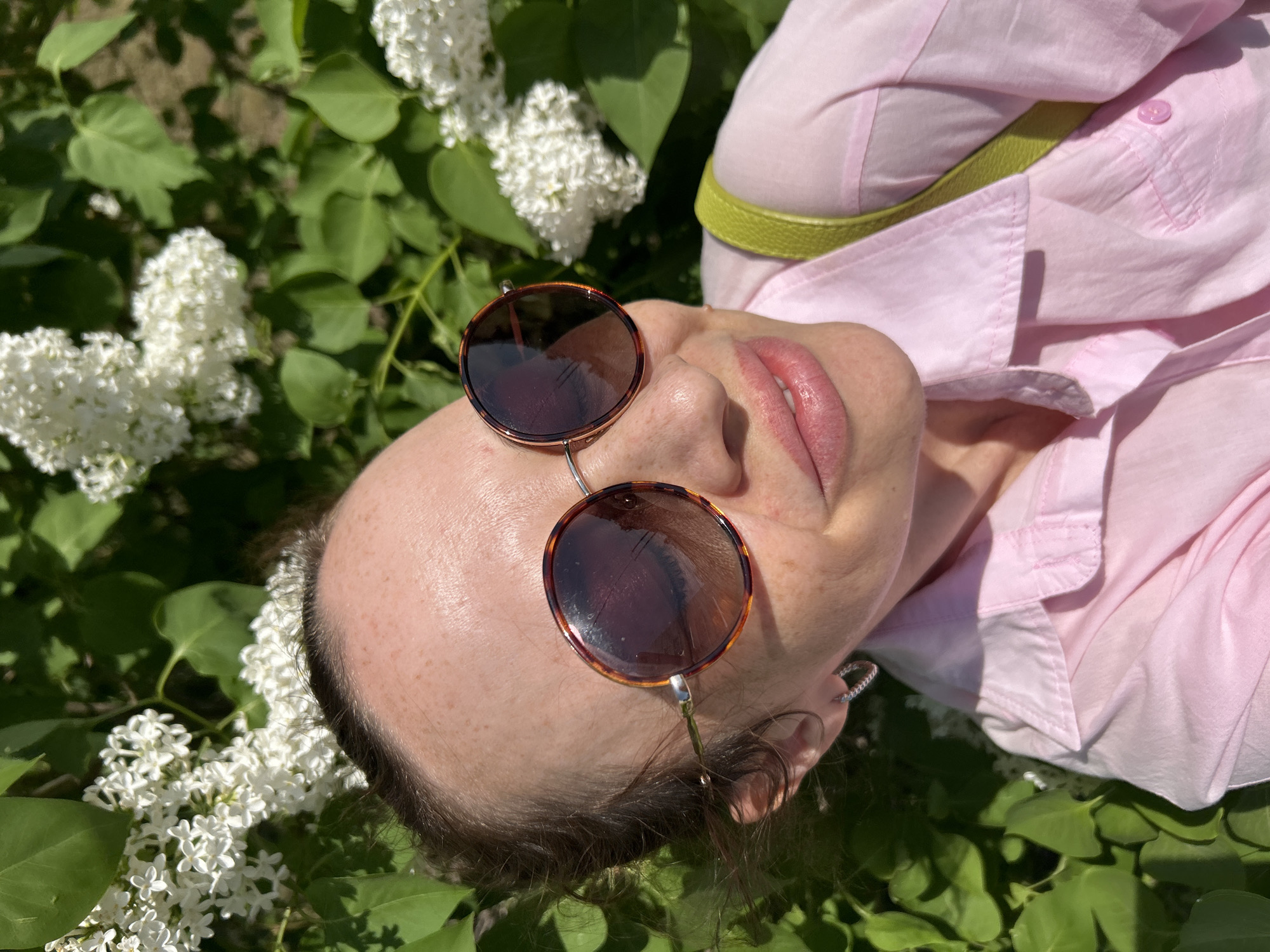 The image shows a person lying on their back in a bed of green leaves and white lilac flowers. The person is wearing a pink shirt with a green strap, and sunglasses are resting on their face. The location is Chaikovsky, Russia.