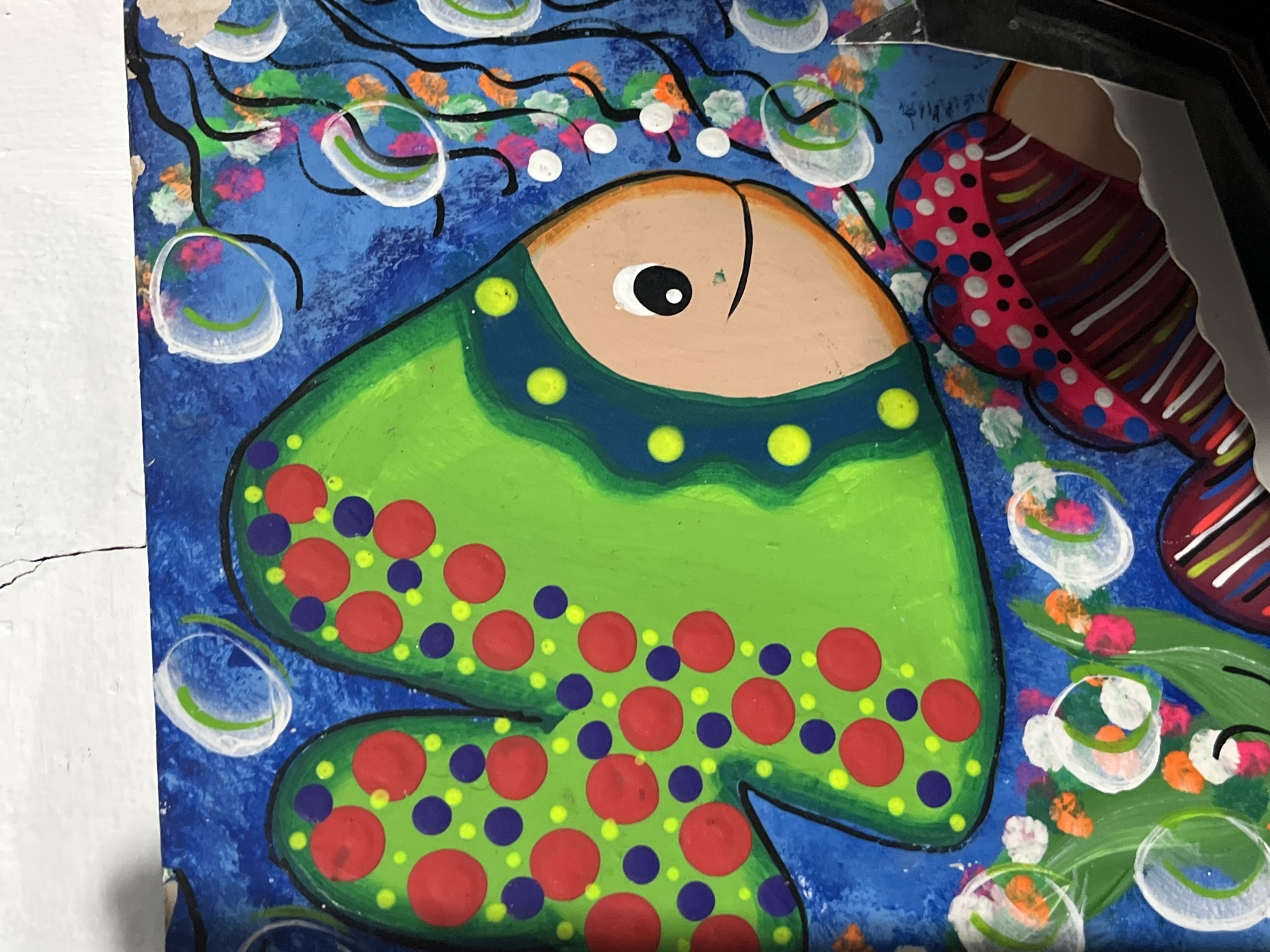 The image shows a section of a painted folk art piece, likely a mural or a painted panel, depicting stylized fish in an underwater scene. The background is a bright blue with painted bubbles and floral or seaweed-like designs. One prominently featured fish is predominantly green, with red and purple polka dots and a darker blue trim with yellow dots around its head. It has a tan-colored head with a single, large black and white eye and a black line for a mouth. Part of another fish, painted with stripes and polka dots in shades of red, blue, and white, is visible on the right side. The artwork is likely from Juan Griego, Venezuela, judging by the style, although this is not explicitly seen within the image itself. The scene seems cheerful and playful, rendered in a naive artistic style.