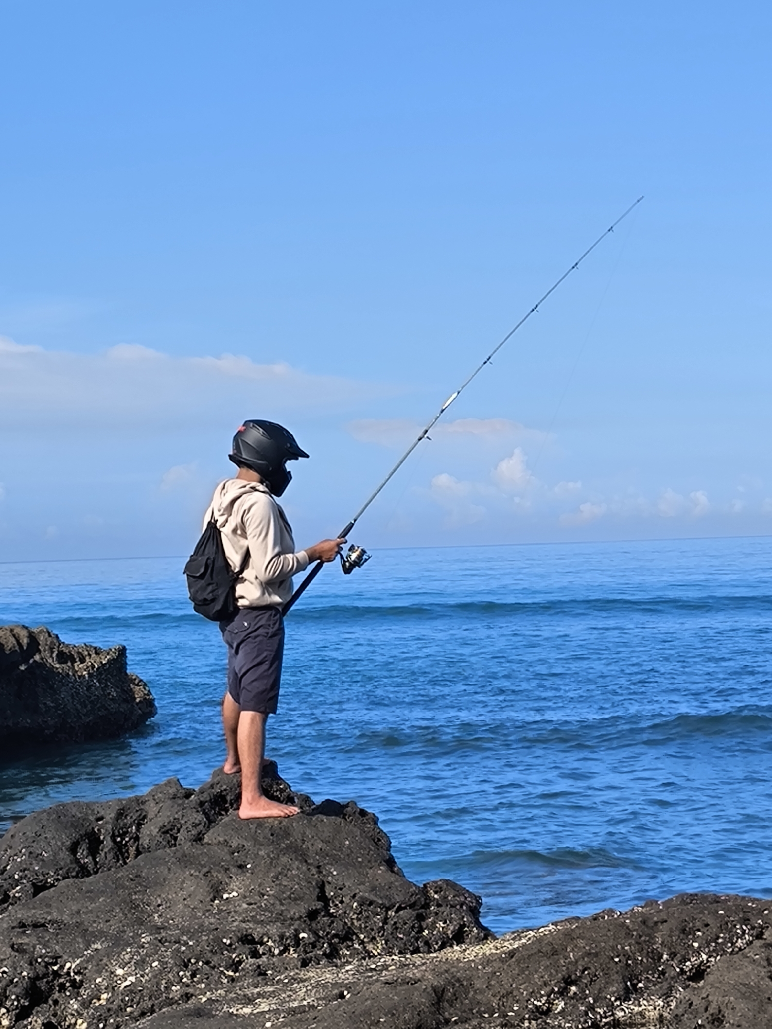 The image depicts a person fishing off a rocky shore in Canggu, Indonesia. The individual, who is wearing a black full-face helmet and a face covering, stands barefoot on a dark, porous rock formation. They are dressed in a light-colored hooded sweatshirt and dark shorts, with a black backpack slung over their shoulder. The person holds a fishing rod with a visible reel, casting a line out into the blue ocean, which stretches to the horizon under a sky with scattered white clouds. Other dark rocks are visible to the left of the person. The location can be presumed to be a coastal area in Canggu, given the rocky shoreline and ocean view.