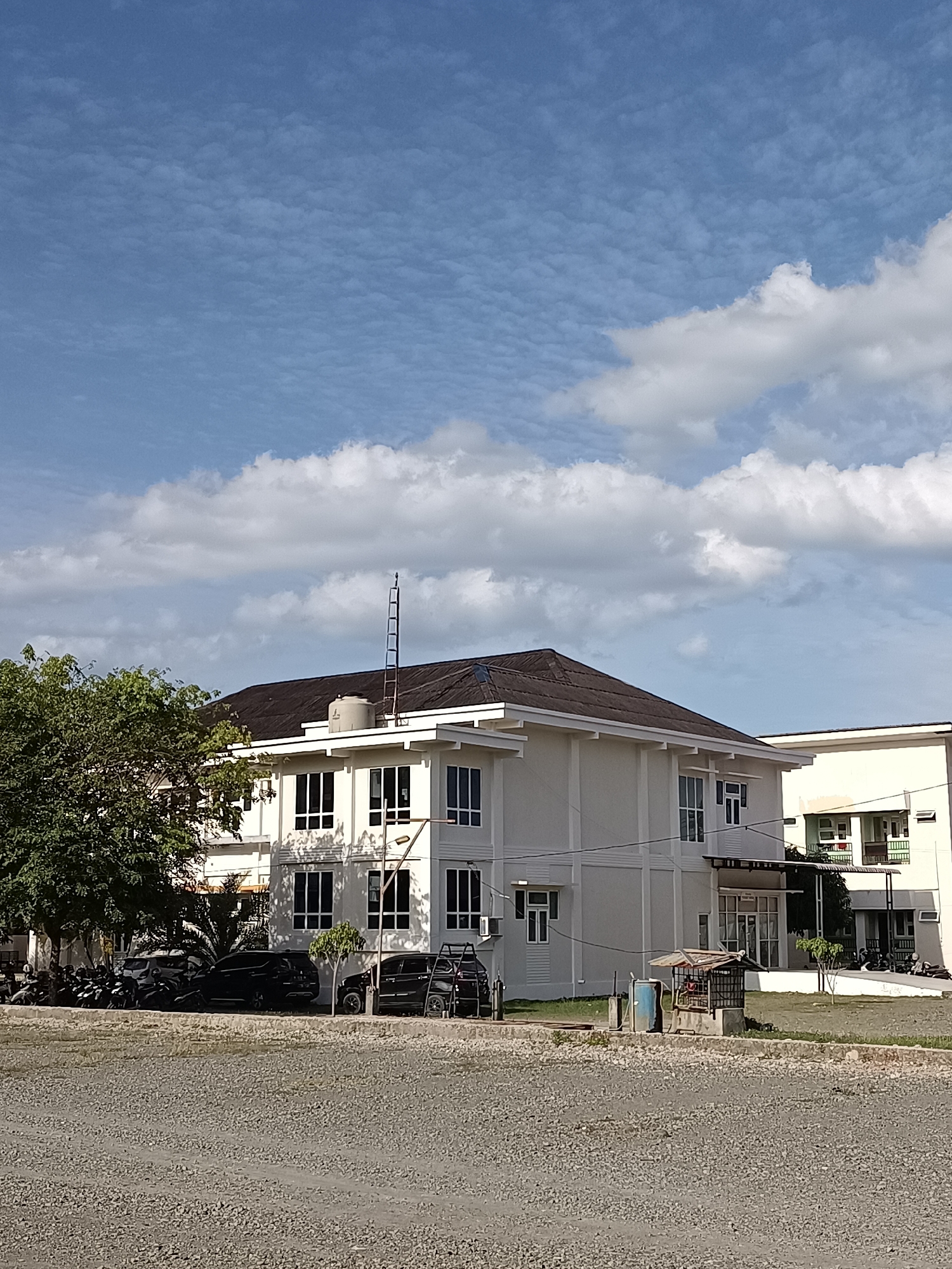 The image depicts a low-rise, light-colored building with a dark, sloped roof, situated in Mesjid Reuntoh, Indonesia. The building has a modern architectural style with numerous windows. In front of the building, several parked cars and motorcycles are visible. A large tree with dense green foliage is on the left side of the frame, and a smaller tree is in front of the building. The ground in the foreground is covered with small, light-colored gravel. The sky above is blue with scattered white clouds. A small, open-sided structure with a corrugated metal roof and a cage-like enclosure stands near the building. A water tank is also visible on the roof of the main building. Another building, lighter in color, is partially visible to the right.