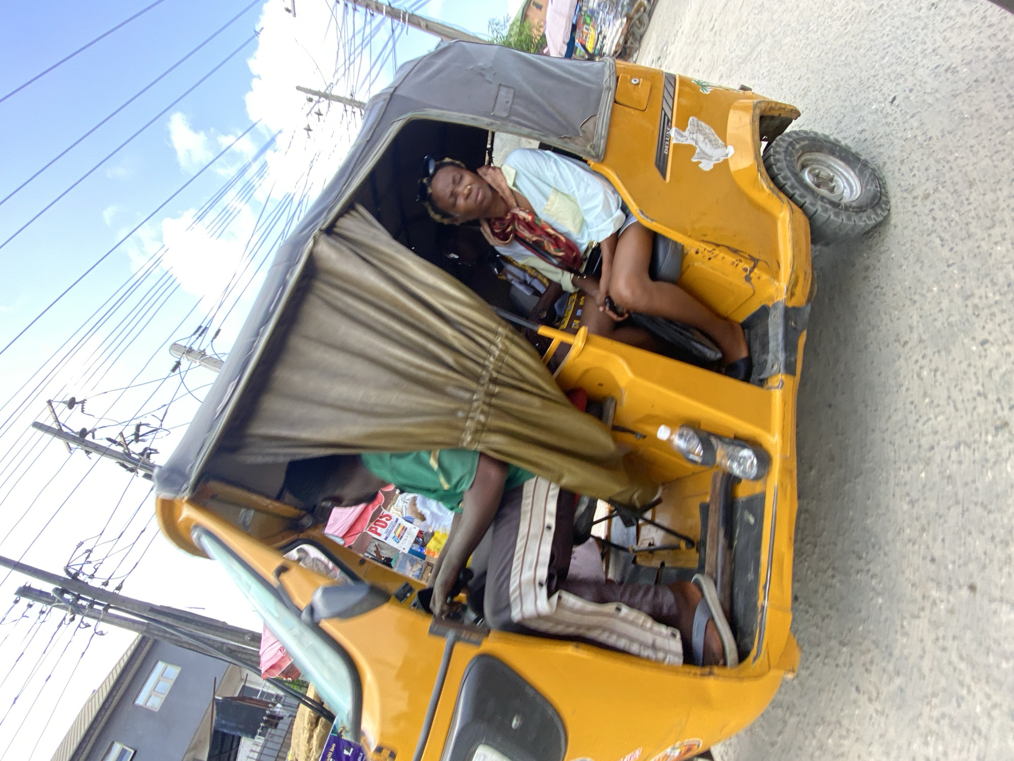 The image, captured in Lekki, Nigeria, shows a bright, daytime street scene centered on a yellow three-wheeled auto-rickshaw, commonly known as a Keke NAPEP. The photograph is oriented vertically, rotated 90 degrees counter-clockwise.
Inside the vehicle, two individuals are present. In the front, the driver, wearing a green shirt and dark pants with white side stripes, is visible with their hands on the controls. Near the driver, several plastic bottles are placed, and an item partially displaying "POS" can be seen. In the passenger area, a woman is seen relaxing or sleeping, with her head tilted back and eyes closed or partially closed. She wears a light-colored long-sleeved shirt, shorts, sunglasses perched on her head, and a patterned red, orange, and white scarf around her neck.
The yellow Keke NAPEP features a dark grey canvas roof and shows some signs of use. The text "DELUXE" is visible on its side, along with a white graphic resembling a turtle. The vehicle is situated on a paved surface, likely asphalt or concrete.
The background is dominated by a clear blue sky with white, fluffy clouds. Numerous power lines stretch across the frame, connected to visible utility poles. Portions of buildings are also discernible in the distance, indicating an urban or semi-urban setting. The scene conveys a moment of daily transportation under sunny weather conditions.