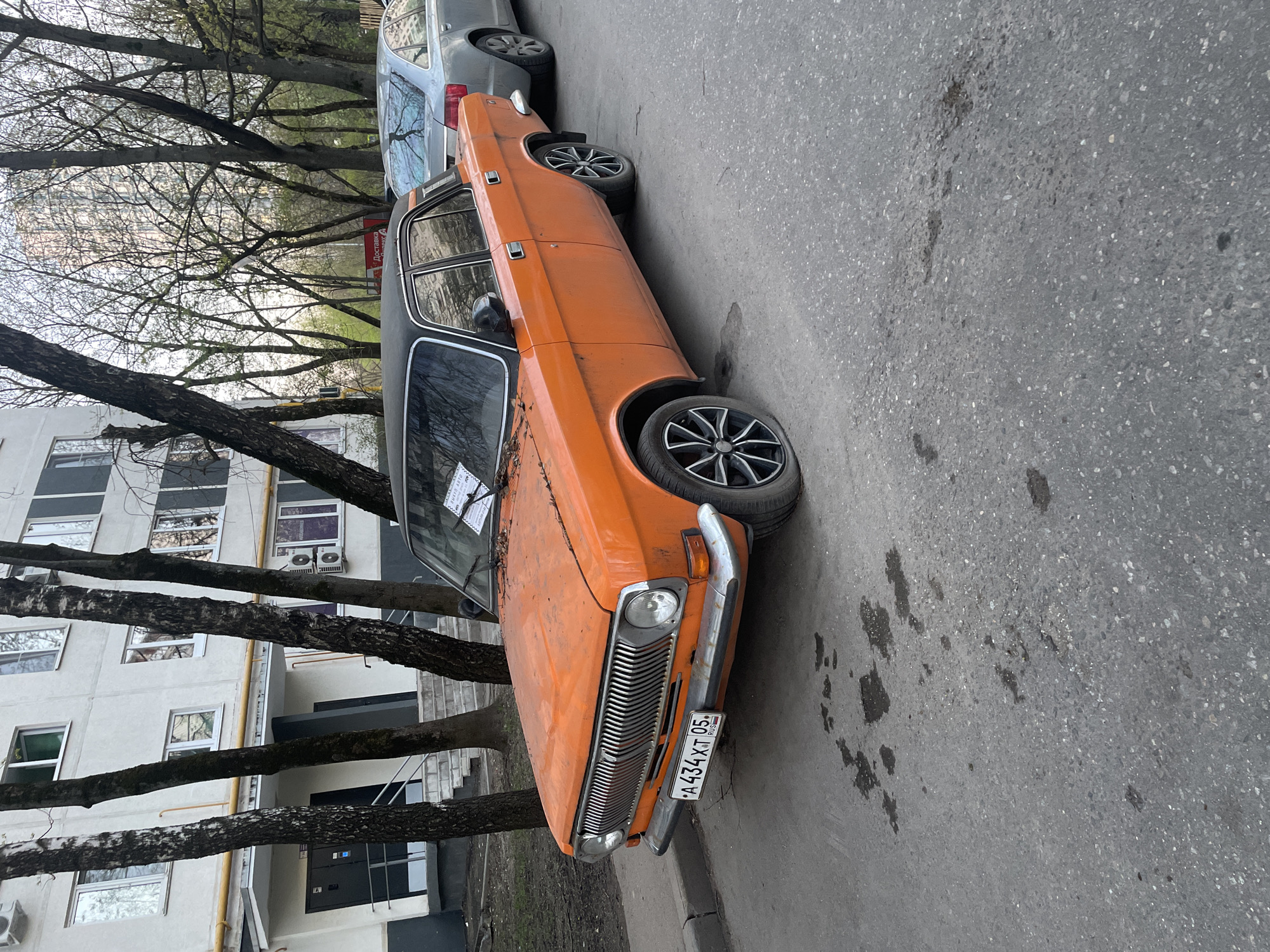 The image depicts an orange, vintage Volga car parked on an asphalt street, likely a residential area in Moscow, Russia. The car is parked in front of an apartment building with bare trees lining the sidewalk. The car's license plate number is A434XT05. Another car, silver in color, is parked behind the Volga. The asphalt shows some dark stains, likely oil or other fluids. The building has several windows and air conditioning units.