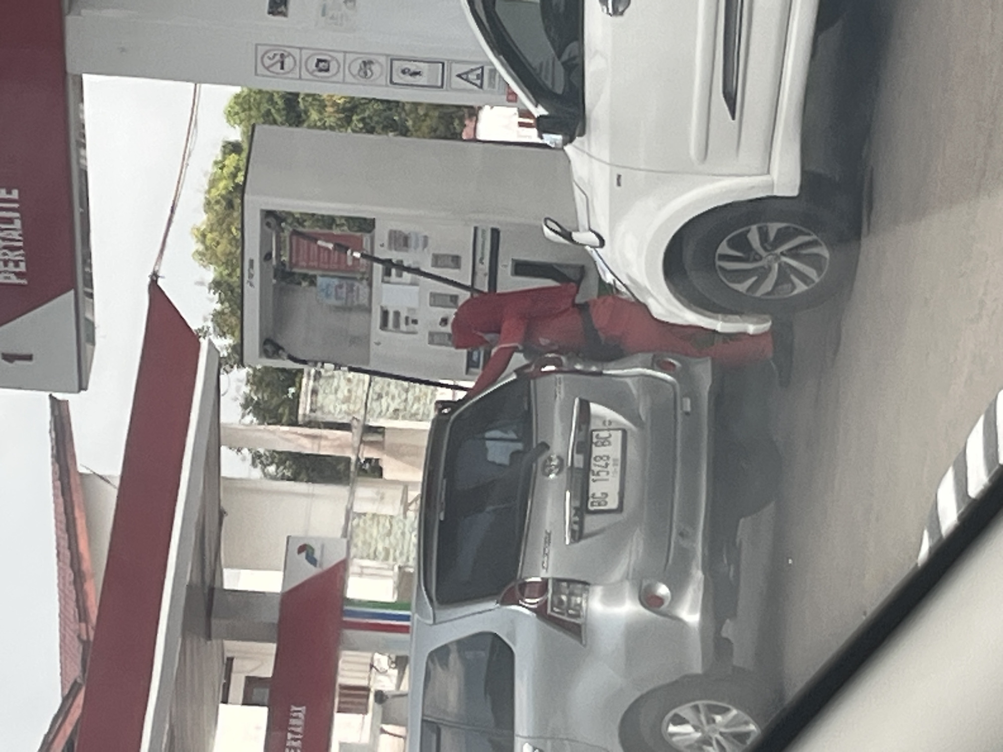 A person wearing a red hijab and red clothing is seen fueling a silver van at a Pertalite gas station in Sekayu, Indonesia. The van has a license plate reading "BG 1548 BC". A white car is parked next to the van. The gas station features red and white awnings and signage. The background shows trees and a cloudy sky.