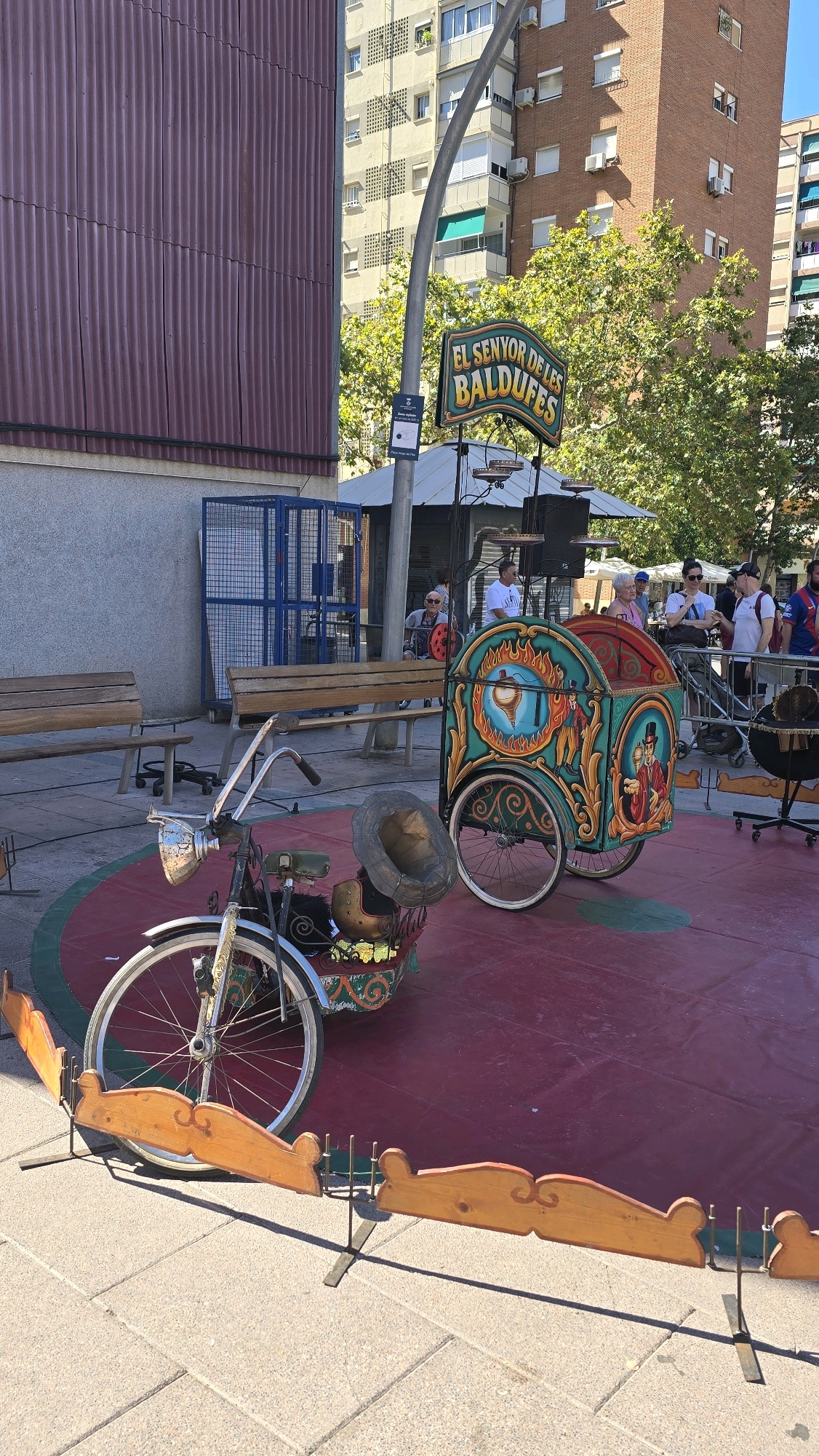 In Cornellà de Llobregat, Spain, a sunny outdoor scene features a red circular stage with a wooden perimeter. A vintage-style, elaborately decorated cart with ornate paintings and two wheels sits on the stage. In front of the cart is a bicycle with a metallic headlight and a hat placed on its handlebars. Two wooden benches are visible behind the stage, and a blue metal cage stands to the left. In the background, a tall brick building and a lighter-colored building with numerous windows dominate the skyline, with trees and a street lamp interspersed. Several people are present in the background, some standing near the cart and others walking. A sign above the cart reads "EL SENYOR DE LES BALDUFES."