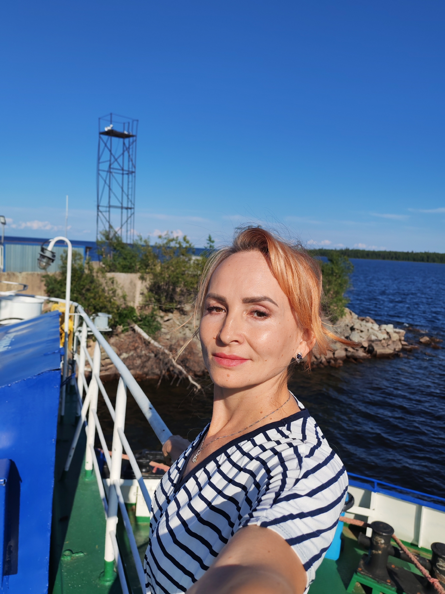 A woman with reddish-blonde hair, wearing a striped dress and a necklace, takes a selfie on the deck of a ferry. She is looking towards the camera with a slight smile. In the background, there is a body of water, a rocky shore with trees, and a metal tower. The sky is clear and blue. The scene appears to be set in Kvartsitny, Russia.