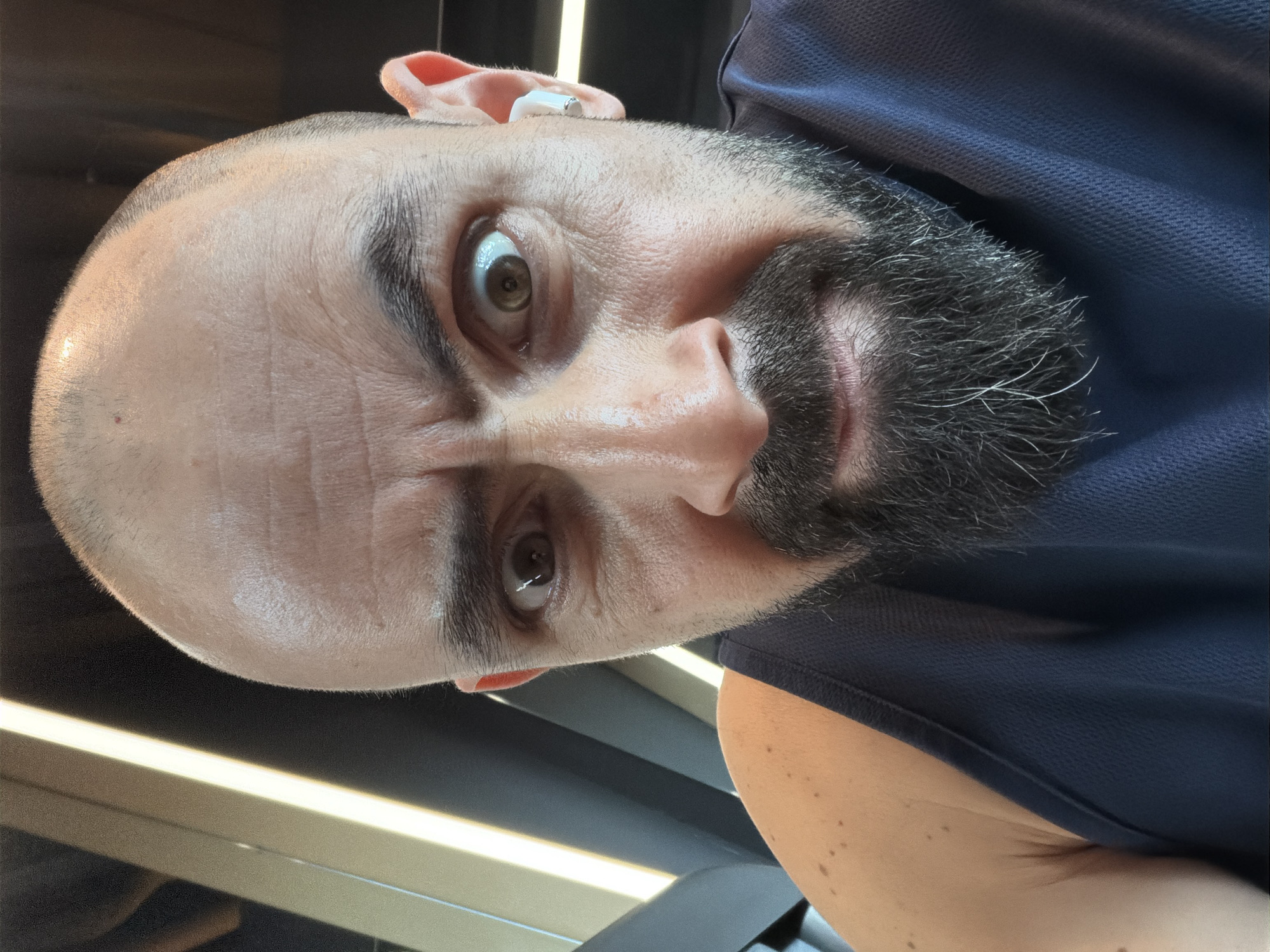 A close-up, front-facing portrait shows an adult male located in Burgas, Bulgaria. The individual has a bald head, dark furrowed eyebrows, and a dark full beard containing visible grey hairs. His eyes are wide open, directed intensely at the camera, with the left iris (viewer's right) showing more sclera. A white wireless earbud is present in his right ear. He is wearing a dark blue, textured sleeveless garment, consistent with athletic wear. The background is out of focus, revealing dark architectural elements and a prominent vertical strip of bright white light on the left, indicative of an indoor, contemporary setting, potentially a gymnasium. The subject's skin exhibits a slight sheen, possibly from perspiration.