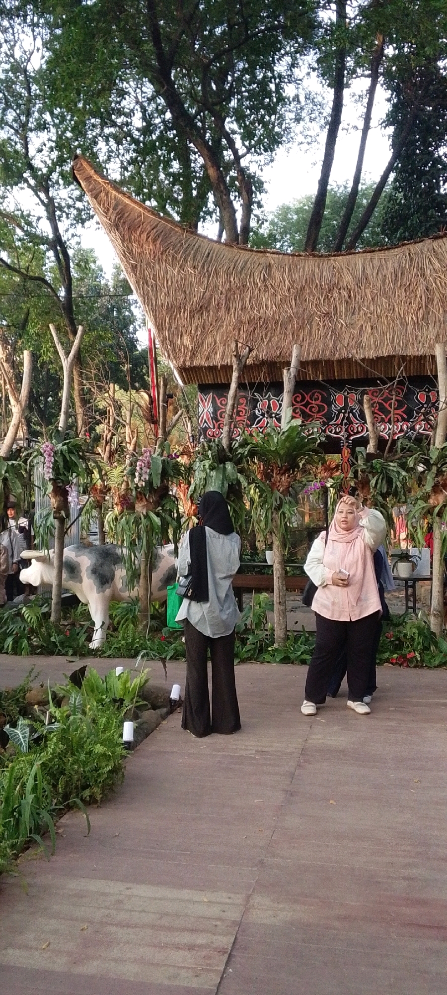 In Central Jakarta, Indonesia, a scene unfolds at what appears to be an outdoor cultural exhibition or garden. A traditional thatched-roof structure with intricate black and red patterned decorations dominates the background. In the foreground, a wooden walkway leads through lush greenery, featuring various plants and flowers. To the left, a life-sized statue of a cow with grey and white patches stands amongst the foliage.
Three women are present. One woman, wearing a grey shirt and black pants, is viewed from the back, her head covered with a black hijab and a small black handbag slung over her shoulder. She stands near the cow statue, facing away from the viewer. Another woman, in a light pink shirt and black pants, is also wearing a hijab and stands facing the viewer. She has one hand raised to her forehead. A third woman, partially visible and wearing a light colored top and dark pants, stands beside her. The lighting suggests it is daytime, possibly late afternoon, with a soft, natural light illuminating the scene. The environment is verdant and appears well-maintained, suggesting a public space designed for leisure and cultural appreciation.