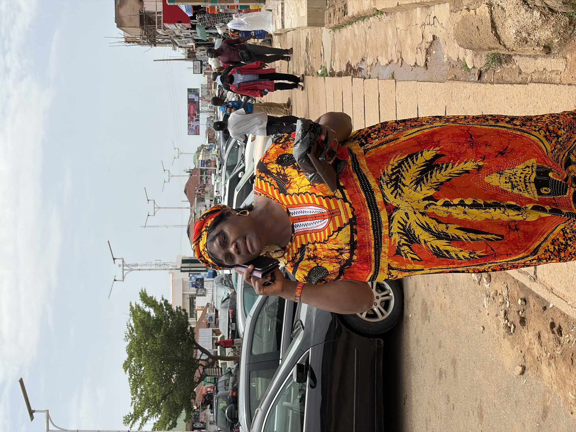 The image shows a street scene in Jos, Nigeria. A woman in the foreground is wearing a vibrantly colored, patterned dress and headscarf, and is holding a mobile phone to her ear, appearing to be in conversation. She is standing on a sidewalk next to a parked car, which is part of a row of vehicles lining the street. In the background, other people are visible walking along the sidewalk and street, and shops and buildings line the road. The scene appears to be during the daytime, with bright, overcast skies. The overall atmosphere suggests a busy urban environment, likely a marketplace or commercial area. There are no other notable emotions or specific events clearly depicted beyond the woman's phone call and the general activity of people in the street.