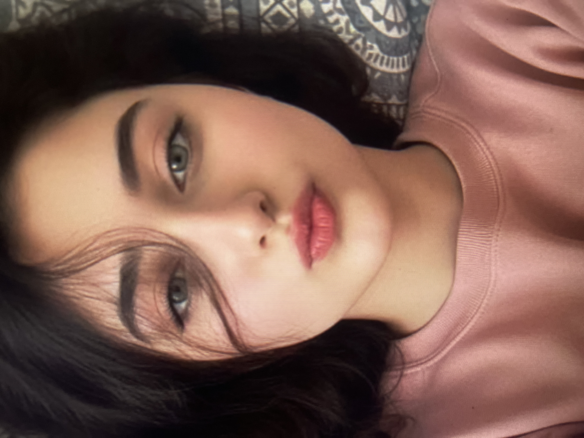 The image shows a close-up, waist-up portrait of a young woman with dark hair and light-colored eyes. She is wearing a pink crewneck sweatshirt. Her face is angled slightly to her left, and her lips are pursed. A patterned fabric is visible behind her on the right side of the frame. There are no visible clues or indicators within the image that would allow for the determination of its location in Qarağandı city administration, Kazakhstan.