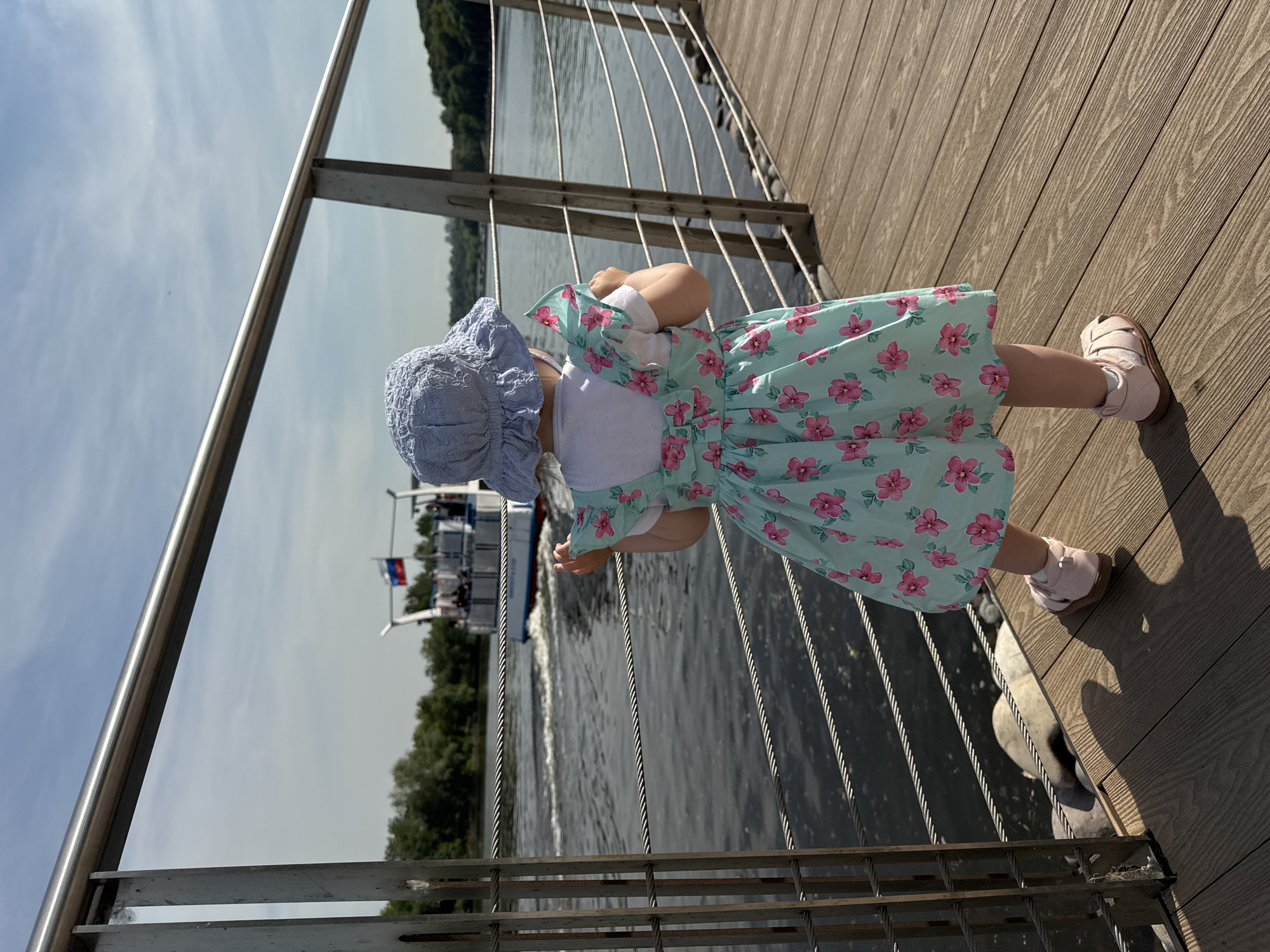 A young child, viewed from behind, stands on a wooden boardwalk or pier, gazing across a river. The child is wearing a light blue sun hat, a white t-shirt, a light blue dress with a pink floral pattern, and light pink sandals. Their hands are positioned near a metal railing, which features a horizontal top bar and multiple horizontal tension cables. In the mid-ground, a white and blue passenger boat with a Russian flag (white, blue, red horizontal stripes) flying at its stern is visible on the dark river, moving from left to right and generating a wake. The opposite bank of the river is densely covered with green trees. The scene is brightly lit by daylight, casting shadows of the child and railing onto the wooden decking. This location is situated along a river in Moscow, Russia.