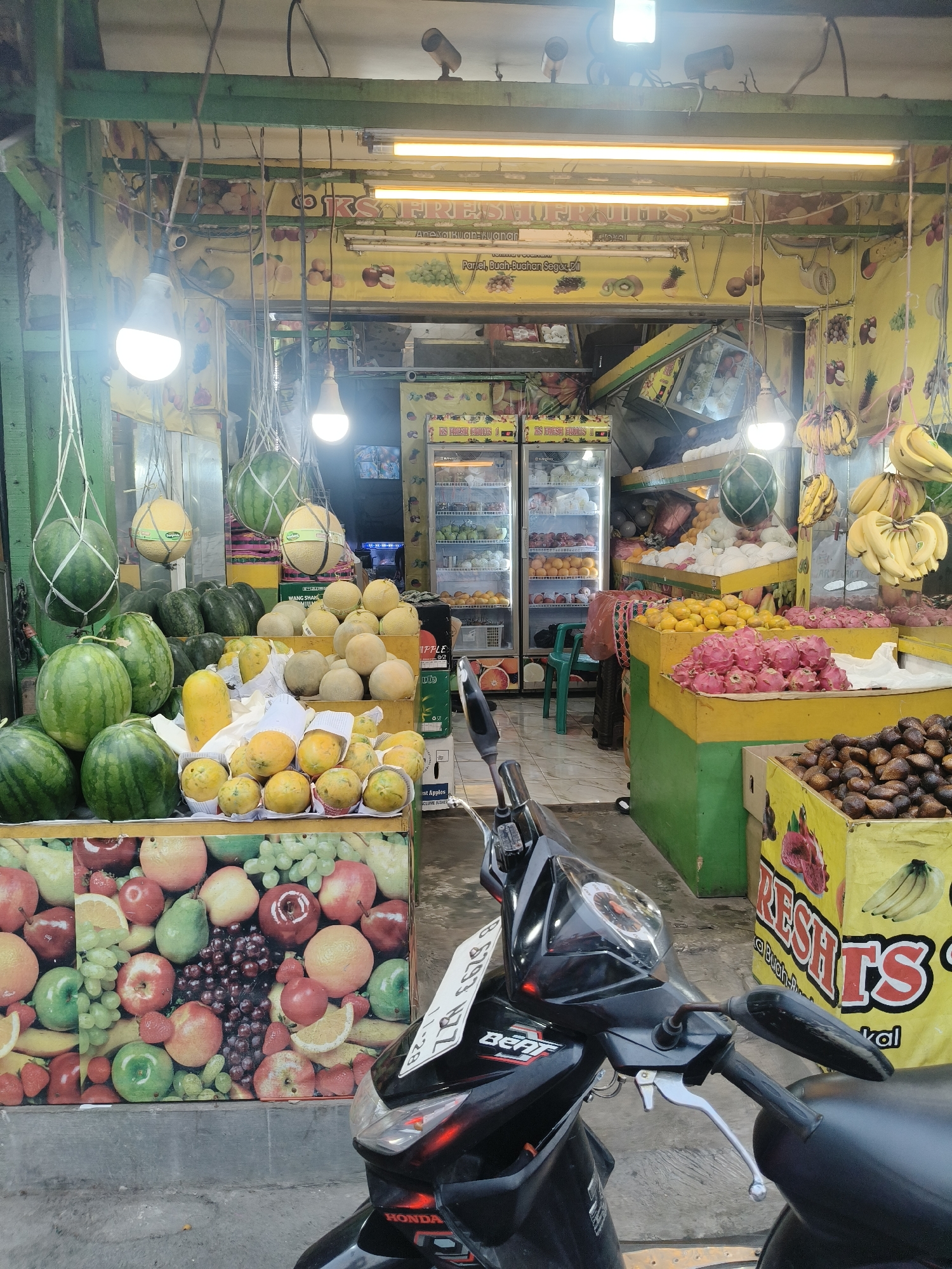 This is a medium shot of a fruit stand in South Tangerang, Indonesia. The fruit stand is open to the street and is well-stocked with a variety of fruits, including watermelons, melons, papayas, oranges, dragon fruit, bananas, and salak (snake fruit). A black Honda Beat scooter is parked in front of the stand, partially obscuring the view of the lower portion of the display. The fruit stand is decorated with colorful murals of fruits. In the background, there are two commercial refrigerators filled with more fruits and possibly other beverages. The lighting appears to be a mix of natural daylight and fluorescent lamps overhead. The overall scene depicts a vibrant and bustling fruit market environment. There are no people visible in the image.