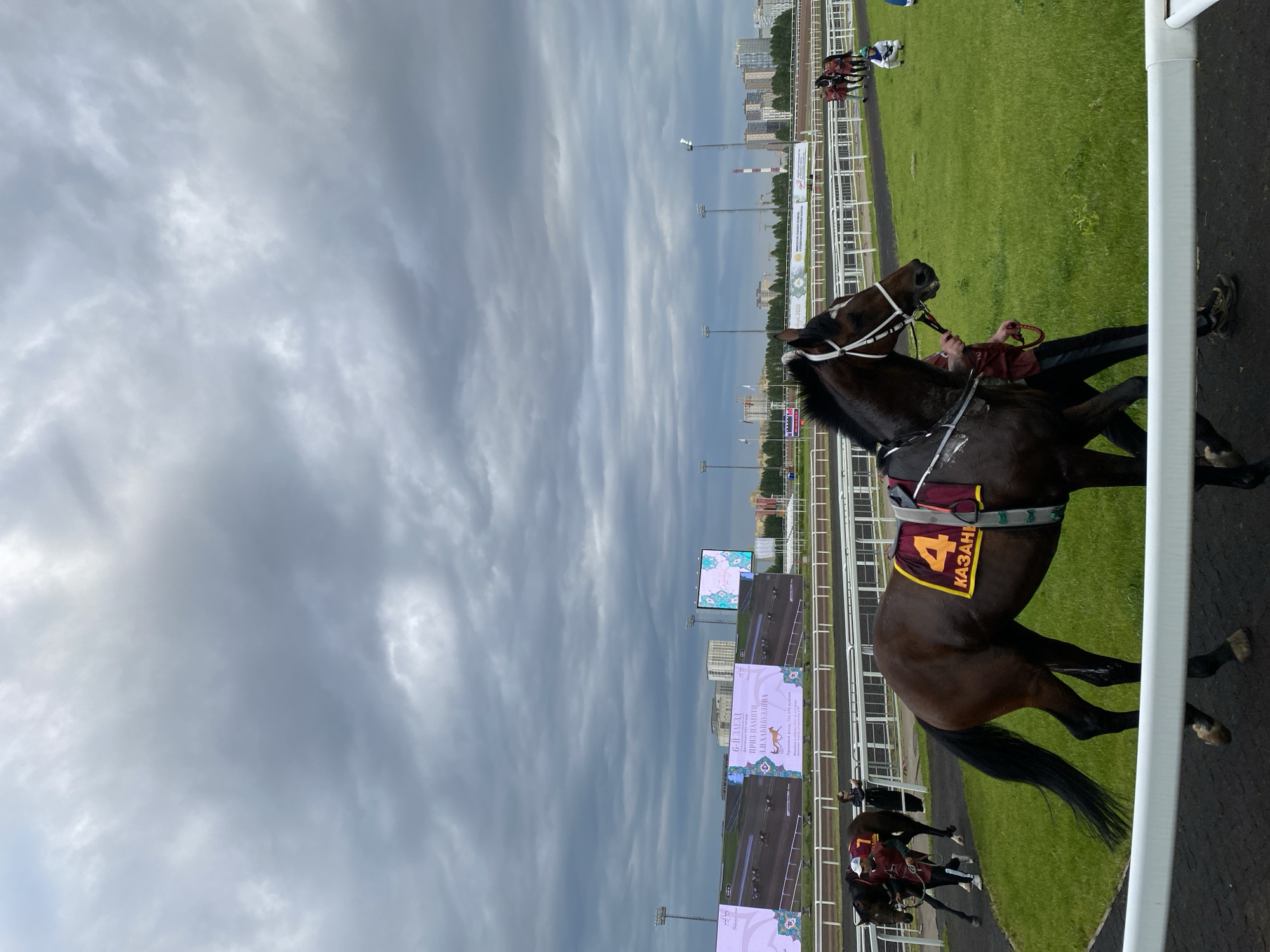 The image depicts a horse racing event at the Kazan Hippodrome, located in Kazan, Russia. The sky is overcast with dramatic gray clouds, suggesting impending rain. In the foreground, a dark brown racehorse, identified by the number "4" on its saddle cloth, is being led by a handler. The saddle cloth also has the word "KAZAN" written on it. The horse is on a dark racing surface, with lush green grass to its right. In the background, a grandstand area is visible with large screens displaying race information and images. Several other horses and handlers are present on the track, along with a person wearing a jockey's silks in the distance, kneeling on the grass. Spectators are not clearly visible. The overall scene is characteristic of an outdoor sporting event under challenging weather conditions.