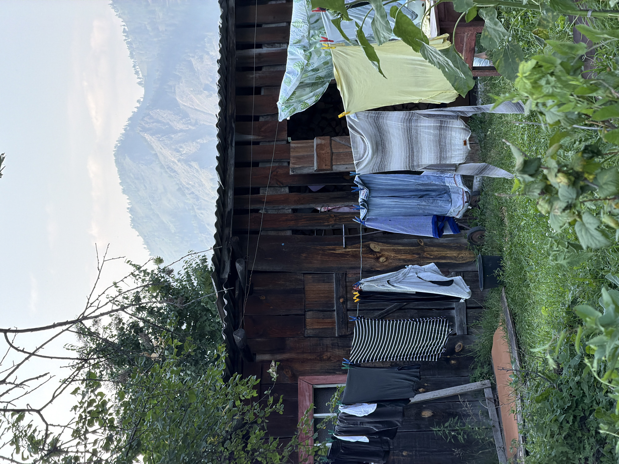 The image depicts an outdoor scene at what appears to be a rural dwelling in Anos, Russia. A wooden structure, possibly a shed or outbuilding, with a corrugated metal roof, is centrally located. Clothes are hanging on a clothesline in front of the structure. The clothes include various garments such as t-shirts, trousers, and what looks like a striped dress or skirt. Some clothes are secured with clothespins. In the foreground, lush green grass and foliage are visible, with some taller plants, possibly sunflowers, to the right. In the background, a hazy view of mountains is seen under a sky with scattered clouds. The overall atmosphere suggests a mundane, everyday scene in a remote, natural setting. There are no people visible in the image.