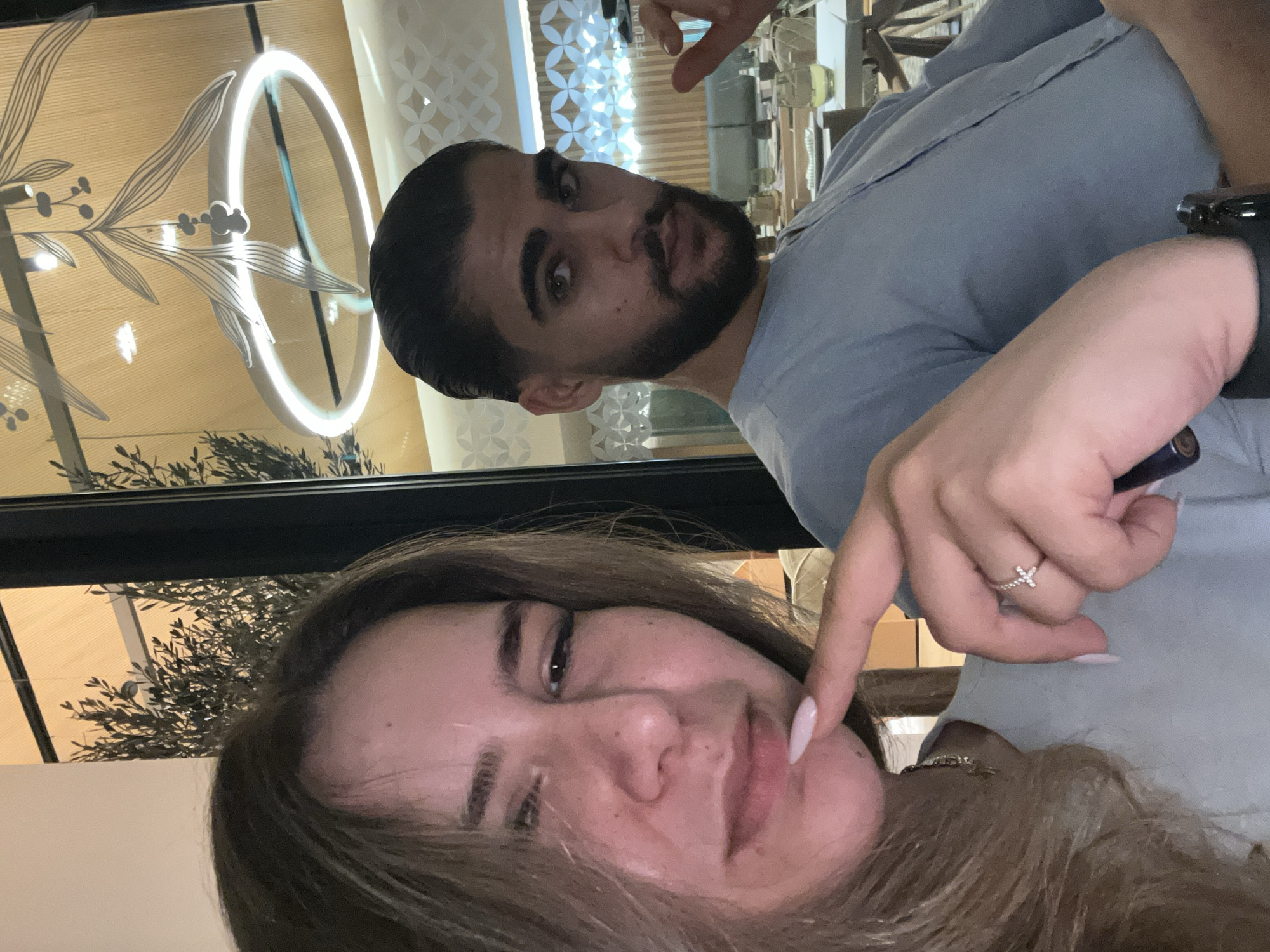 A selfie captured at dusk inside a restaurant in Nicosia, Cyprus shows a young woman and a young man. The woman, positioned in the foreground on the left, has long, wavy brown hair and is wearing a light blue shirt. She is looking at the camera with a slight smirk and has her left index finger touching her lower lip. She is wearing a delicate ring with a cross design on her left hand. The man is on the right, also wearing a light blue shirt. He has dark, slicked-back hair and a beard, and he is looking directly at the camera with a serious expression. In the background, through a large window, is the dimly lit interior of the restaurant, with modern decor including a circular pendant light with a branch motif. The setting suggests a contemporary dining establishment.