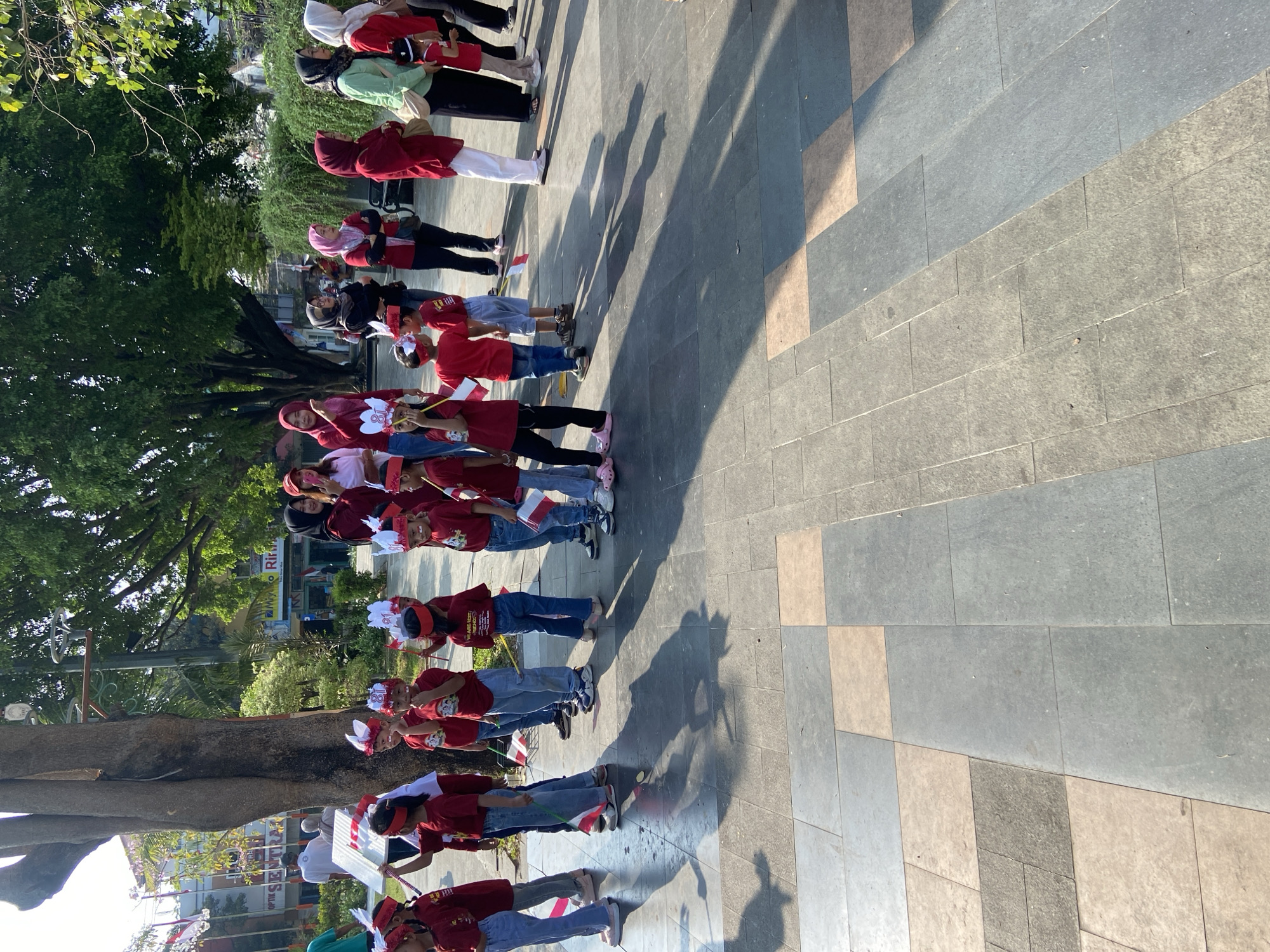 In Kecamatan Pati, Indonesia, a group of children, dressed in matching maroon t-shirts and red and white headbands, stand in a line. Several of the children are holding small red and white flags, likely symbolizing the Indonesian flag. The children appear to be participating in a parade or event, as evidenced by their organized formation and the festive headwear. Behind the children, adults in a variety of attire observe the scene. The background reveals a paved area with large square tiles, bordered by a tree with lush green foliage and a building with signage. The overall atmosphere suggests a community gathering or celebration.