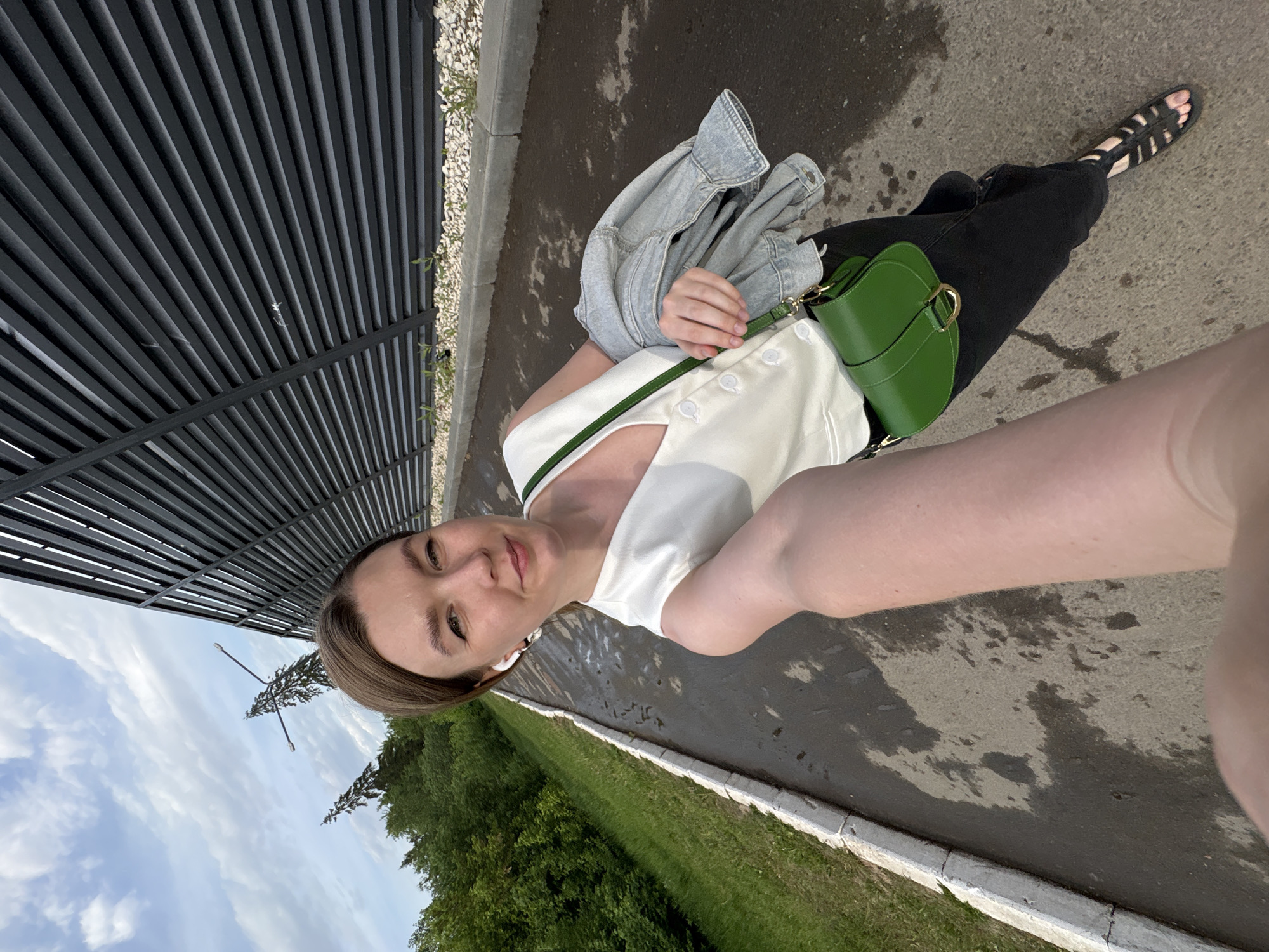 A waist-up, self-portrait photograph shows a young woman in Izhevsk, Russia. The woman is fair-skinned with light brown hair pulled back, wearing a white sleeveless top with buttons, black jeans, and black sandals. A light denim jacket is draped over her left arm, and she carries a small, green, half-moon-shaped bag. She wears what appears to be a white wireless earbud in her left ear.
The photograph is taken from a low angle, looking upwards. The background includes a section of a black metal slatted structure, possibly a bus shelter or a fence. To the right, there is a paved sidewalk and beyond that, a strip of green grass and trees. The sky with clouds is visible in the upper left corner. The woman is holding the camera out and tilted slightly downward toward her face.
