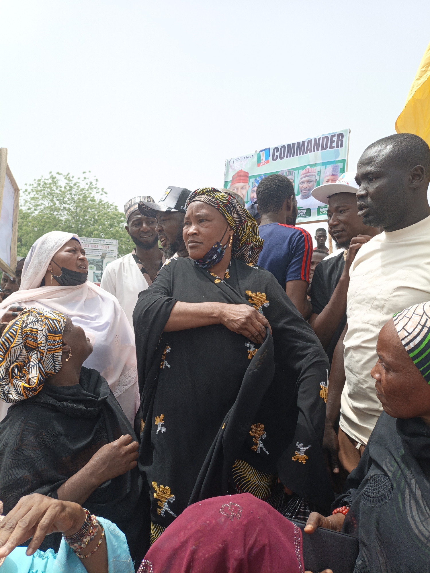 This outdoor photograph captures a daytime gathering of people, predominantly adults, under a bright, clear sky in Giginyu, Nigeria. The scene suggests a political event, characterized by a large banner visible in the background. The banner prominently displays "COMMANDER" in bold lettering, alongside "Vote First APC," and features headshots of several men, including those identified as "Haruna Jowa" and "Murtala Gwa," presumably political figures or candidates.
The foreground is filled with a diverse group of individuals. Several women are visible, many wearing traditional attire such as hijabs and patterned headscarves. One woman in the center wears a black garment adorned with distinctive yellow and orange embroidered patterns and a black face mask. To her left, another woman in a light-colored hijab and a black face mask looks upwards. Other women wear dark clothing and colorful head wraps, some accessorized with bracelets and earrings.
Among the men, some wear traditional caps and tunics, while others are dressed in more casual attire like t-shirts and baseball caps. They are standing in close proximity, engaged in observation or conversation. A partial yellow flag or fabric is visible on the far right of the frame, and a wooden frame of another sign is on the far left. The overall atmosphere indicates a public assembly or rally.