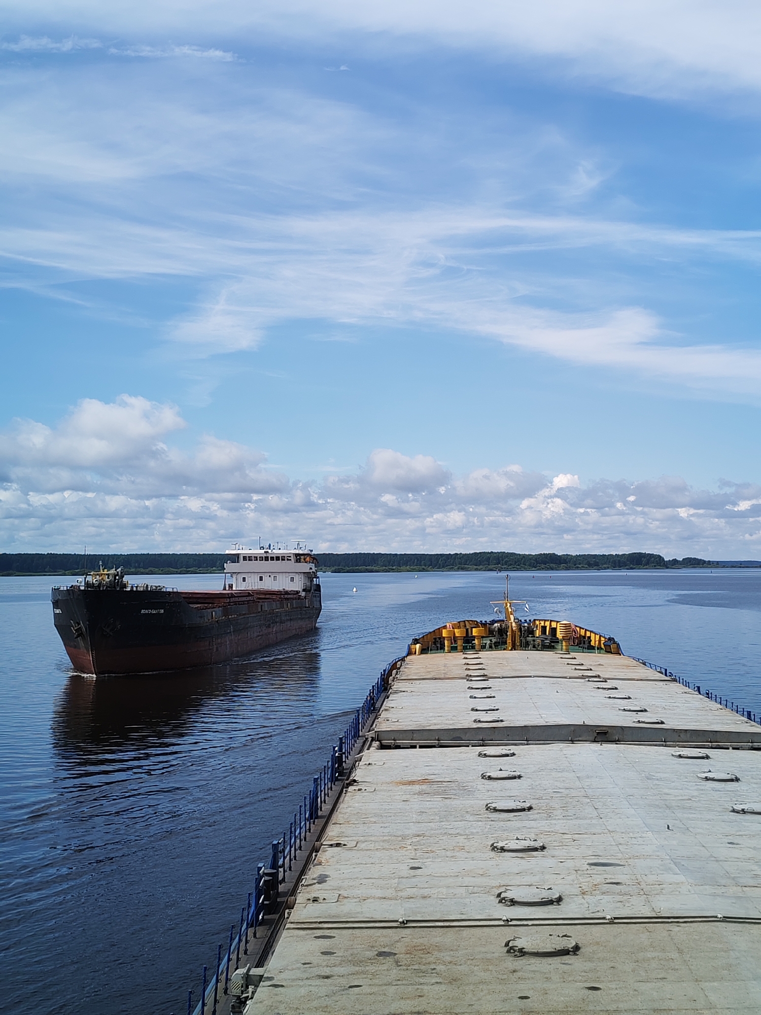 An elevated, wide-angle shot captures two barges on a wide, calm river under a partly cloudy blue sky. The barge in the foreground is moving away from the viewer, its long, flat deck covered in numerous hatches. A yellow mast with a flag is visible at the stern. The barge in the background, a dark-hulled vessel with a white superstructure, is moving towards the viewer from the left. Its nameplate appears to read "ВОЛГО-БАЛТ 195". The riverbanks are lined with dense green trees. The scene is likely the Sheksna River in Cherepovets, Russia, as it is a major waterway in the region and the style of the barges is consistent with river traffic in that area. There are no people visible in the image. The overall impression is one of peaceful, industrial maritime activity.