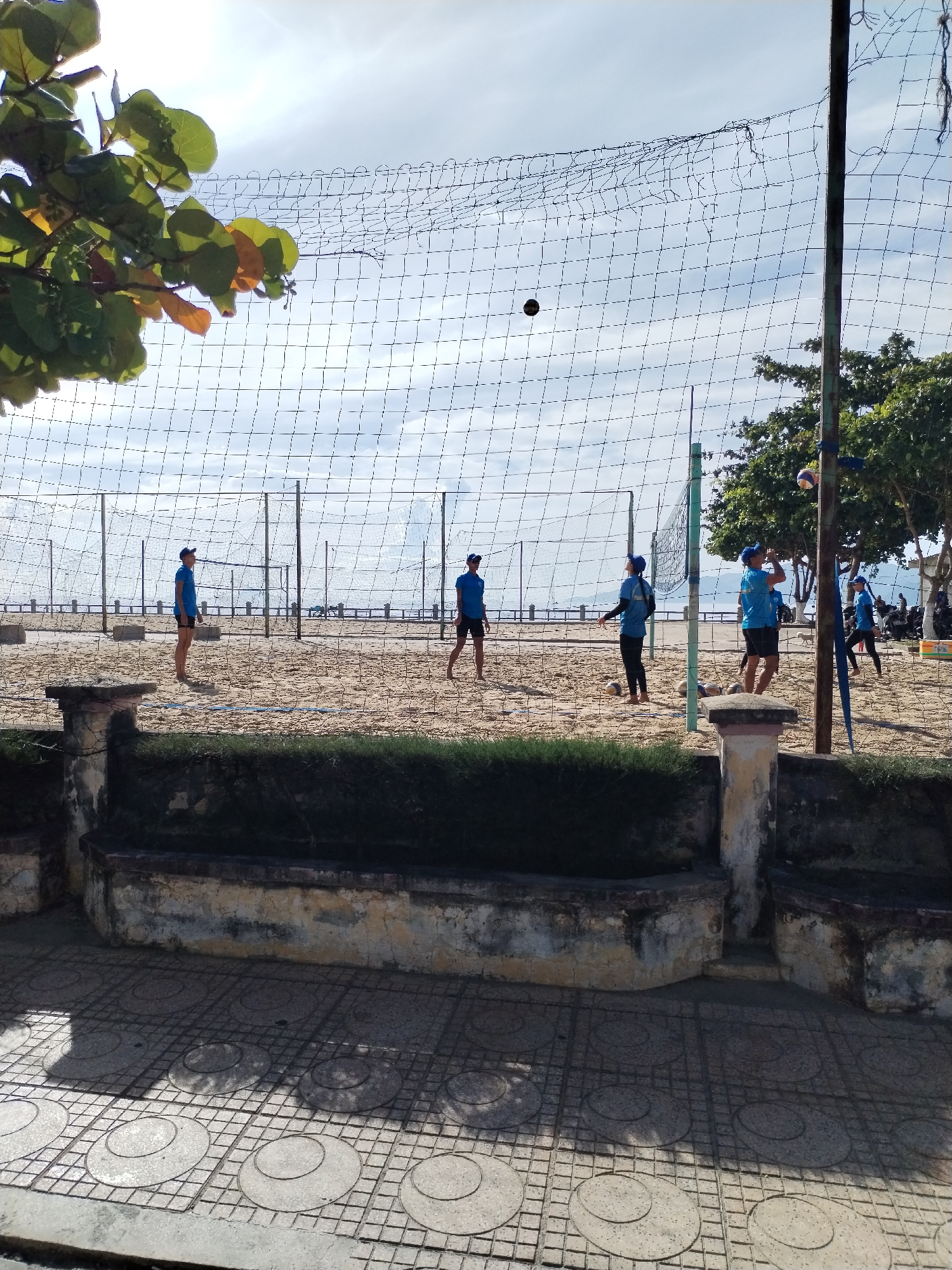 A sunlit outdoor scene depicts people playing volleyball on a sandy beach in Nha Trang Ward, Vietnam. Five individuals wearing matching blue shirts and dark shorts are seen on the beach, positioned around a volleyball net. One person is in the action of hitting a ball. Other volleyballs are scattered on the sand. The net is stretched between wooden poles. In the background, a concrete barrier, trees, and a cloudy sky are visible. In the foreground is a weathered concrete barrier with vegetation growing behind it, and a patterned sidewalk.