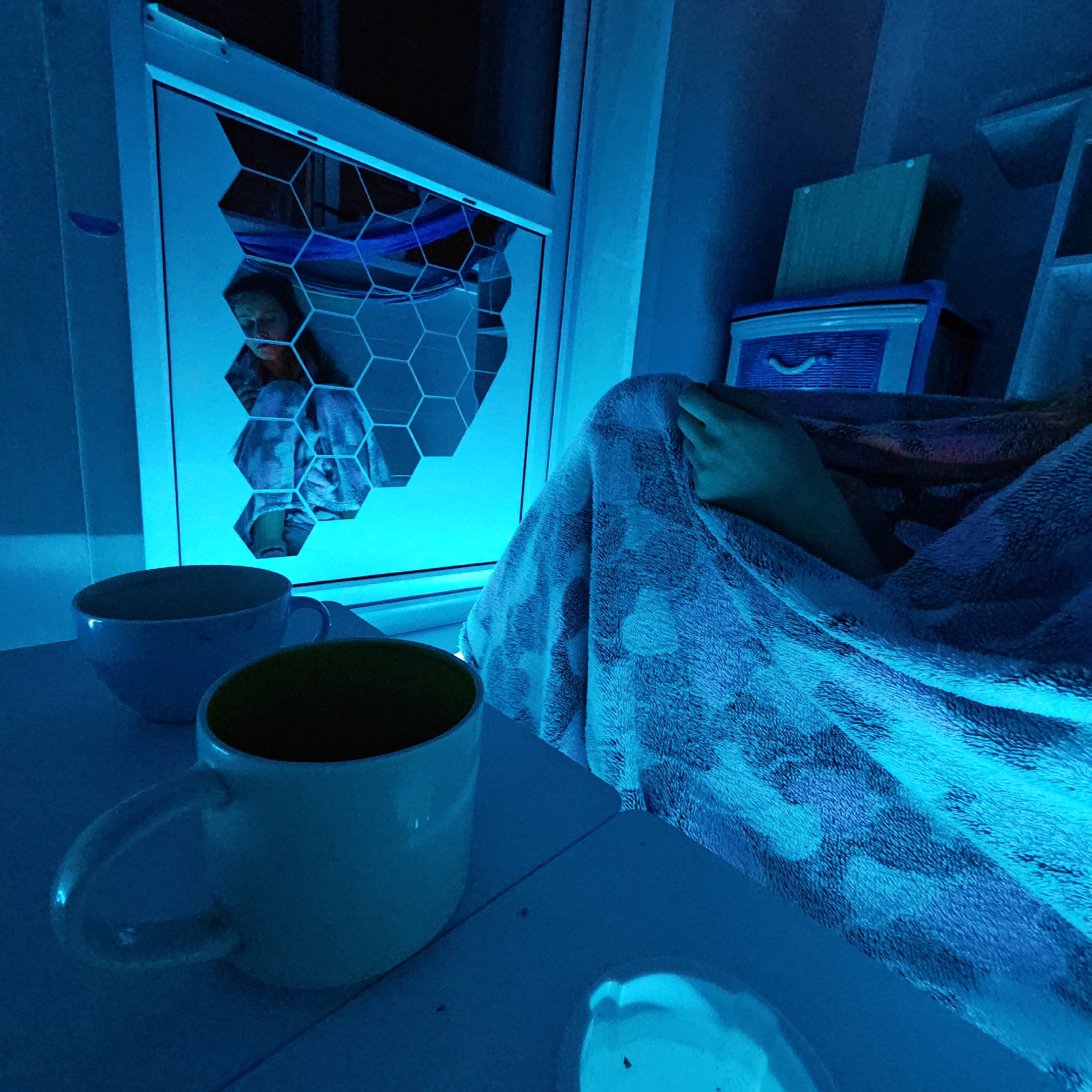 A low-angle, medium shot shows a dimly lit room with a person under a blanket, next to a window with a geometric pattern, possibly located in Rostov, Russia, based on the style of the window. Two mugs, one partially visible, sit on a surface near the window. The room's lighting has a strong blue tint, affecting the colors of the objects and surfaces. A shelf with a storage container is visible in the background. A person, presumably female, is slightly visible through the window, reflected in the geometric design.