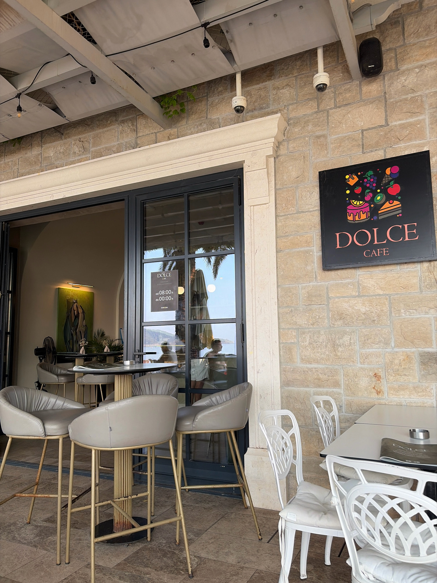 An outdoor seating area of a cafe is pictured in Rafailovići, Montenegro. A sign on the stone wall reads "DOLCE CAFE" with colorful fruit and cake graphics. To the left, the entrance to the cafe features large glass doors. Inside, a painting of horses hangs on the wall. Outside, there are high-top tables with grey leather chairs and round tables with gold accents, as well as white metal tables with white chairs. Some people are visible inside the cafe, and one person is seated at an outdoor table in the distance. The area appears to be near a body of water, as indicated by the palm trees and the view in the background. The weather is overcast, and the lighting suggests it is daytime.