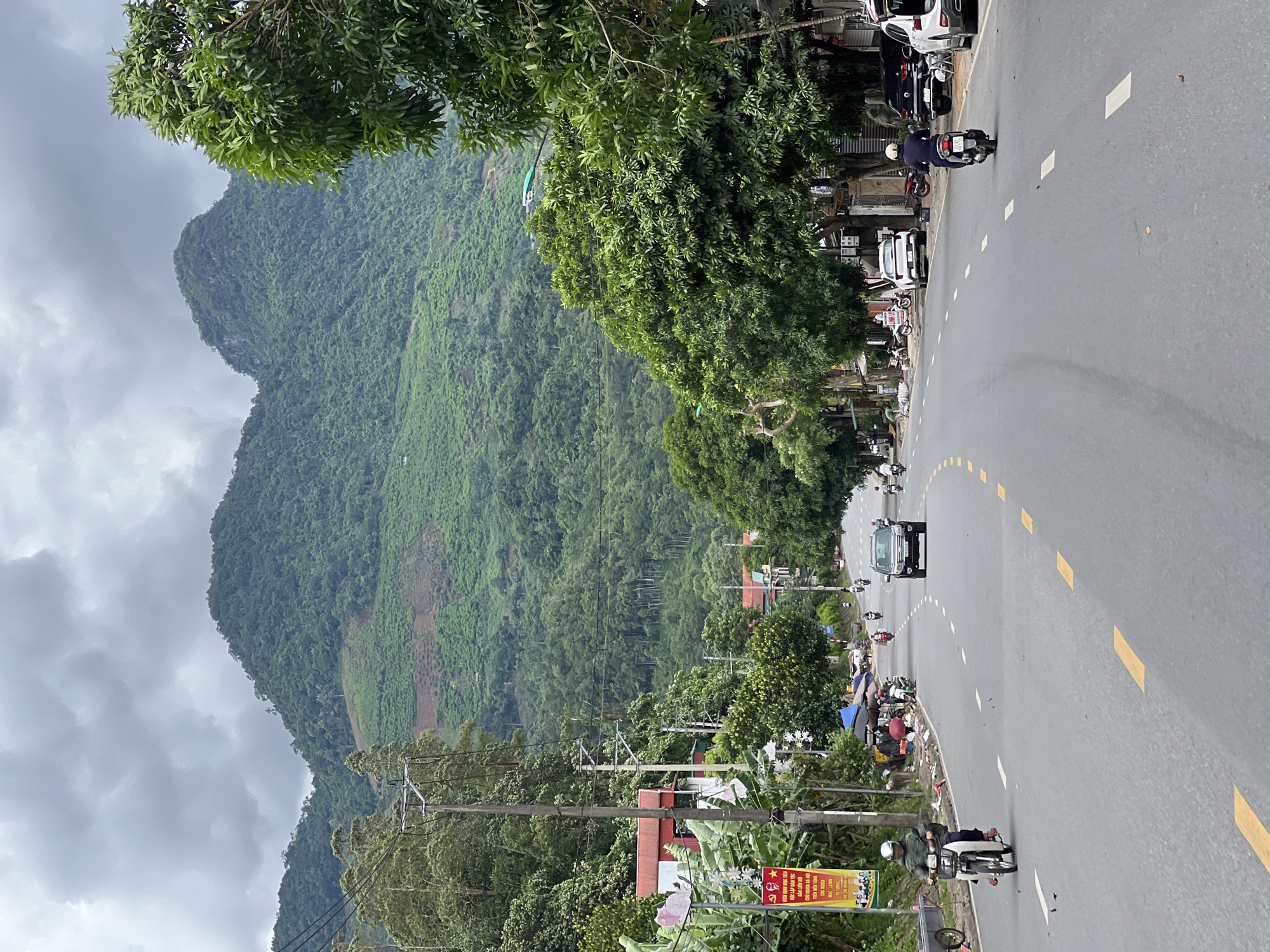 The image depicts a street scene in Hà Giang 2 Ward, Vietnam, characterized by a paved road lined with trees and buildings. The road runs through a valley with lush, green, heavily forested mountains dominating the background. The sky is overcast with heavy clouds.
On the left side of the road, there are a few buildings with red roofs, interspersed with banana trees and a large banner displaying Vietnamese text and images. Electrical poles and wires are visible along the roadside. Several parked motorcycles and scooters are present. A person on a white scooter, wearing a green jacket and helmet, is riding towards the viewer. Further down the road on the left, there are some stalls or makeshift shops covered with umbrellas.
The road itself is a two-lane asphalt surface with yellow and white lane markings. Several vehicles are present on the road. A dark-colored SUV is driving away from the viewer. On the right side of the road, there are more trees and buildings. A person on a black scooter, wearing a dark jacket and helmet, is riding away from the viewer. Parked cars, including a white sedan and a black SUV, are visible along the right curb.
The overall impression is a moderately busy street in a semi-rural or suburban area, nestled within a mountainous landscape under cloudy weather.