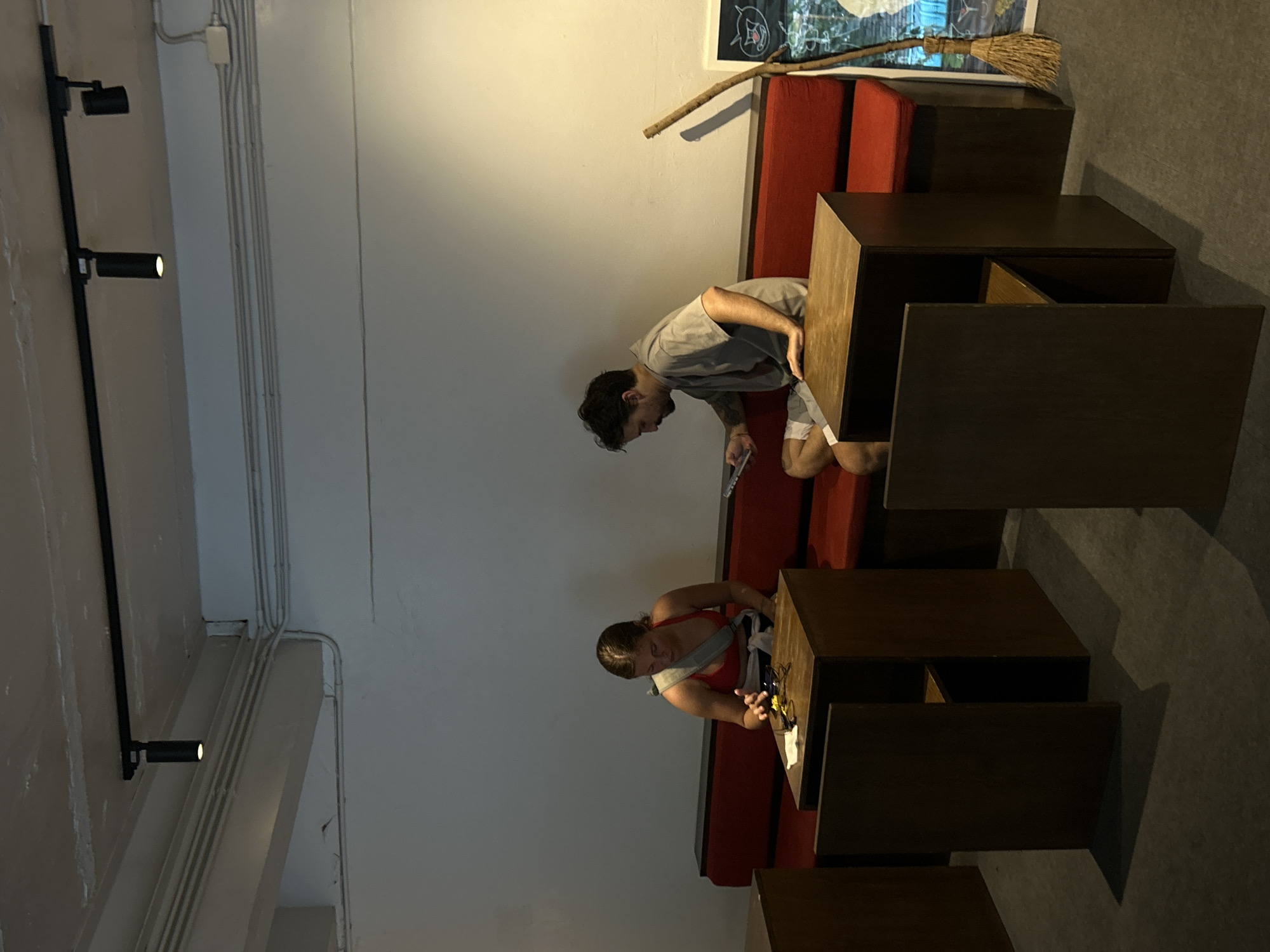 The media file shows two individuals in a dimly lit indoor setting that appears to be a cafe or a communal workspace. The scene is oriented sideways, with the ceiling running vertically on the left and the floor horizontally at the bottom.
The subjects are a man and a woman. The man, wearing a grey t-shirt and shorts, is bent over a wooden table, looking down at something in his hands, possibly a phone or a device. He has dark hair and a mustache. The woman, wearing a red top and grey straps, is also focused on a table, her hands engaged with an object on the surface. She has dark hair tied back.
Several wooden tables of varying sizes and configurations are visible, some appearing as modular units. A red bench is situated behind the man. In the background, a broom with a straw head and a wooden handle leans against the wall above the red bench. The walls are painted in light and darker tones, with electrical conduits and junction boxes visible on the lighter section. Two black spotlights are mounted on a vertical pole on the left side of the frame, with one emitting light.
The overall atmosphere suggests a casual, perhaps late afternoon or evening setting, given the indoor lighting. There is no visible text in the image. The location context of Chiang Mai City Municipality, Thailand, is provided, but no specific visual cues within the image definitively confirm this location.