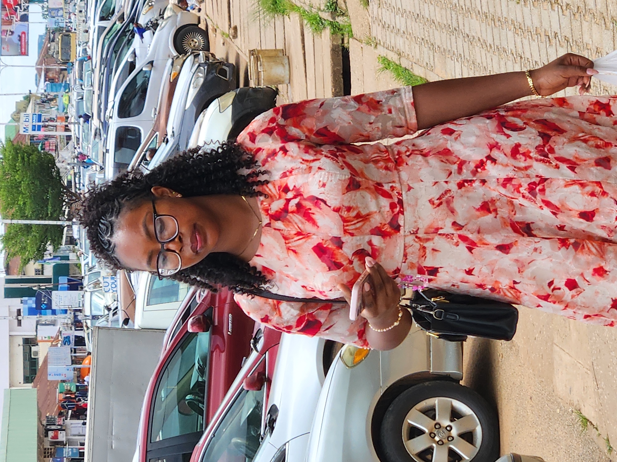 A woman stands on a sidewalk next to a row of parked cars in an urban environment. She is wearing glasses, a floral-print dress, and a gold bracelet. She is holding a smartphone in her right hand and a white plastic bag in her left hand. Her hair is styled in braids. The background shows more parked cars, buildings, and street signs. The ground is paved with concrete and some patches of grass. The lighting suggests it is daytime, possibly overcast.