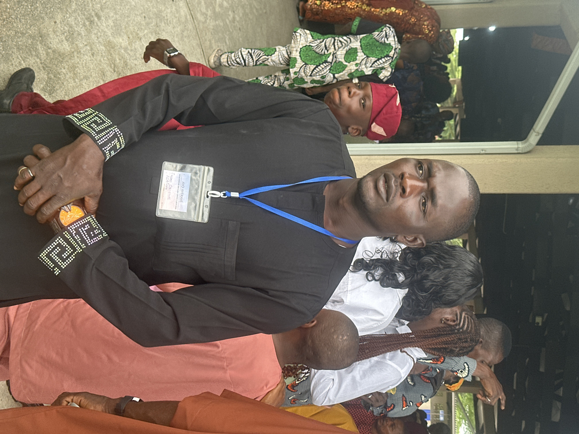 A medium shot captures a group of people gathered outdoors, possibly at an event. In the foreground, a man wearing a black kaftan with intricate white and green beaded cuffs and a blue lanyard around his neck is the primary focus. He holds a small, round, orange object in his right hand and wears a ring on his left ring finger. His left hand is partially visible, with a smartwatch on his wrist. Behind him, a young boy in a red cap and a white and green patterned outfit is looking to his left, with his head tilted. To the right of the main subject, other individuals are visible, including a person in a peach-colored garment and another in an orange garment. In the background, more people are present, and some are pointing or gesturing. The environment appears to be a paved area with a covered structure overhead, suggesting a public gathering space. The overall scene has a vibrant, communal feel.