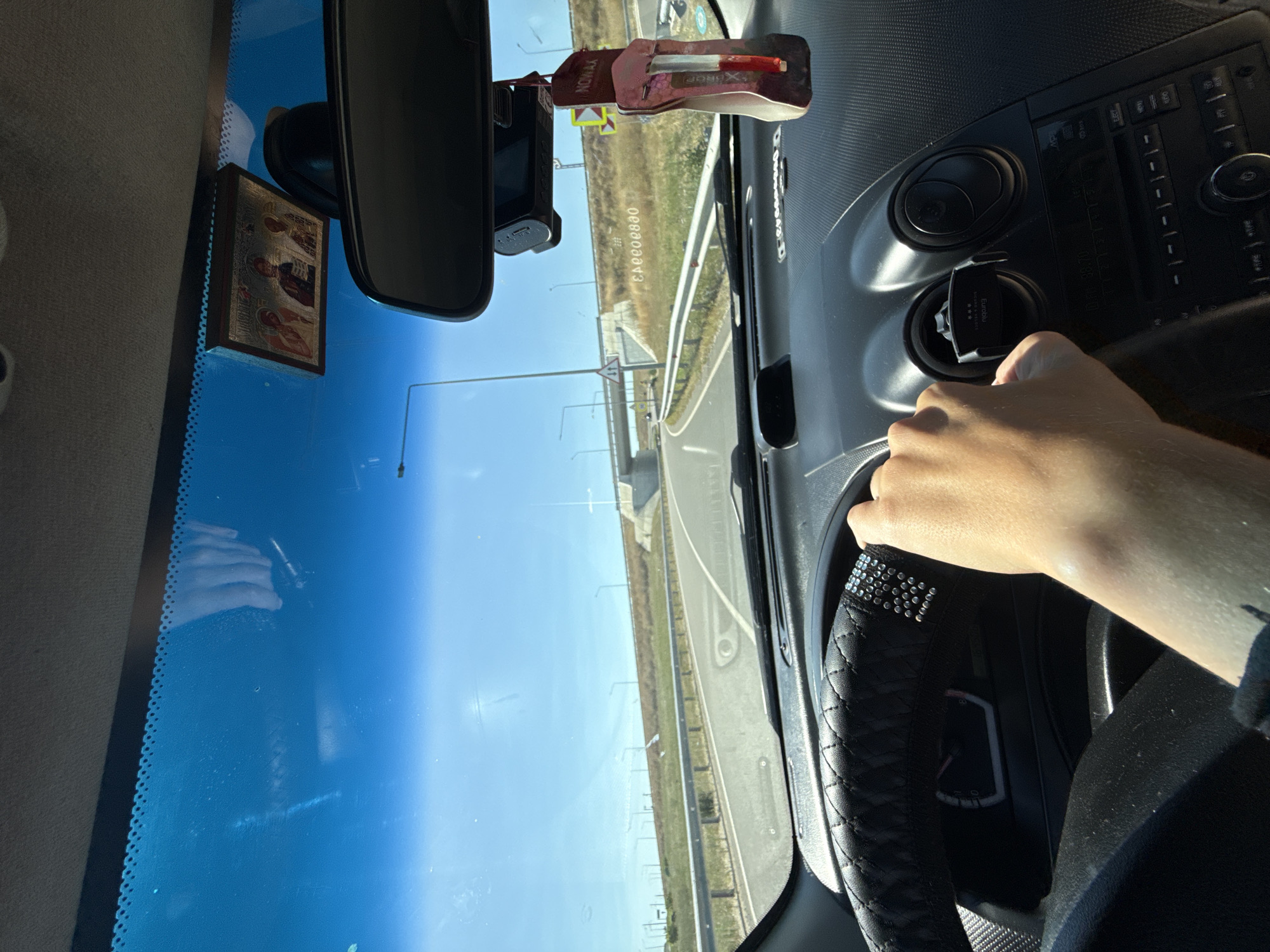 The image shows the interior of a car as seen from the driver's perspective, driving on a sunny day. The dashboard is visible, along with the steering wheel partially covered by a hand. A car air freshener hangs from the rearview mirror, and a small religious icon is attached to the windshield. Outside the car, a highway with multiple lanes is visible, with a metal guardrail and grassy verges. The sky is clear and blue. The location is identified as Cioc-Maidan, Moldova.