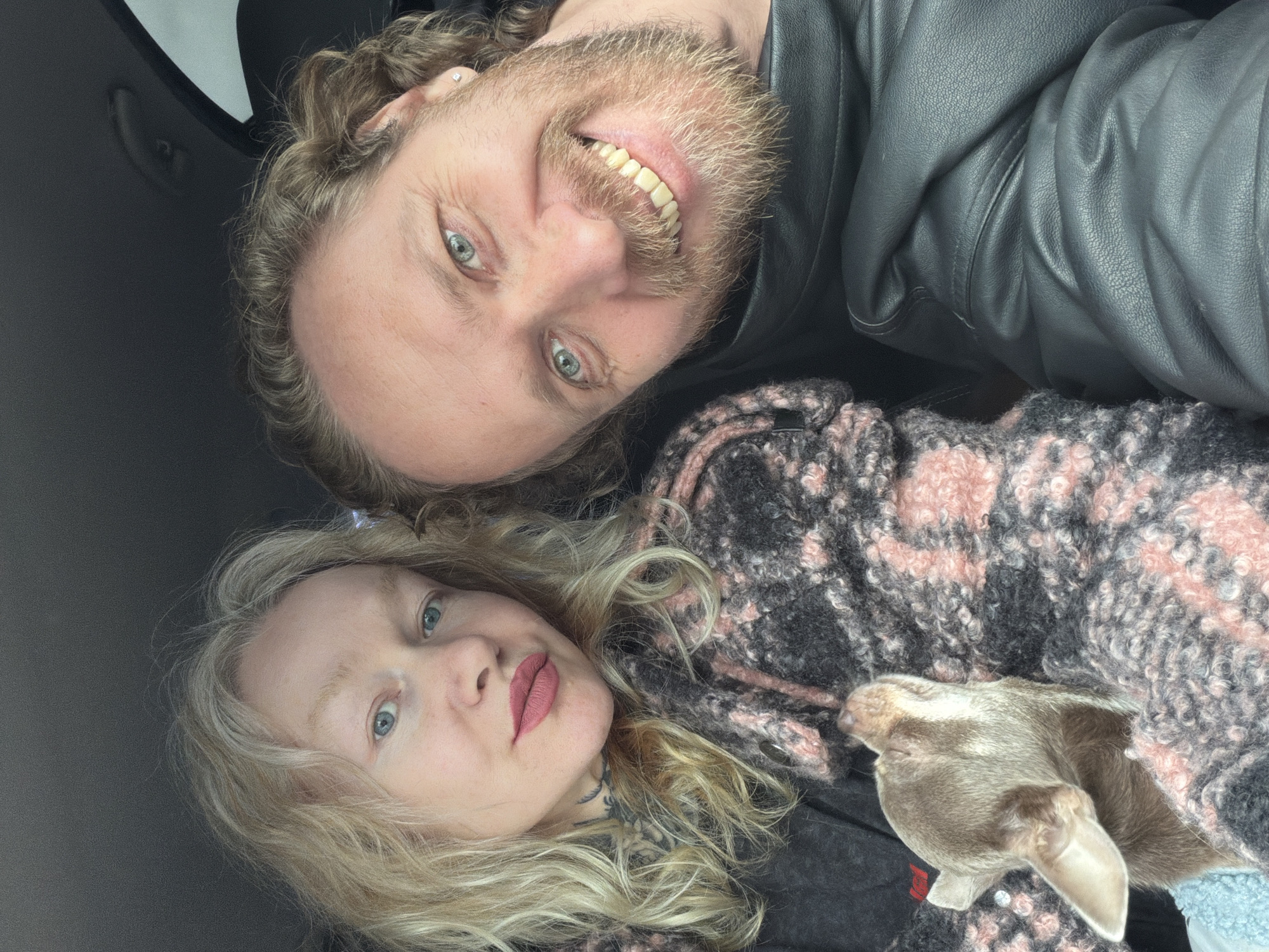 A photograph shows a man and a woman in what appears to be a vehicle. The man is wearing a black leather jacket and has a beard, blonde hair, and green eyes. He is smiling. The woman has blonde, wavy hair, light skin, and is wearing a patterned sweater. A small brown dog is nestled in the sweater next to her. The location in the city of Torosovo, Russia, cannot be determined.