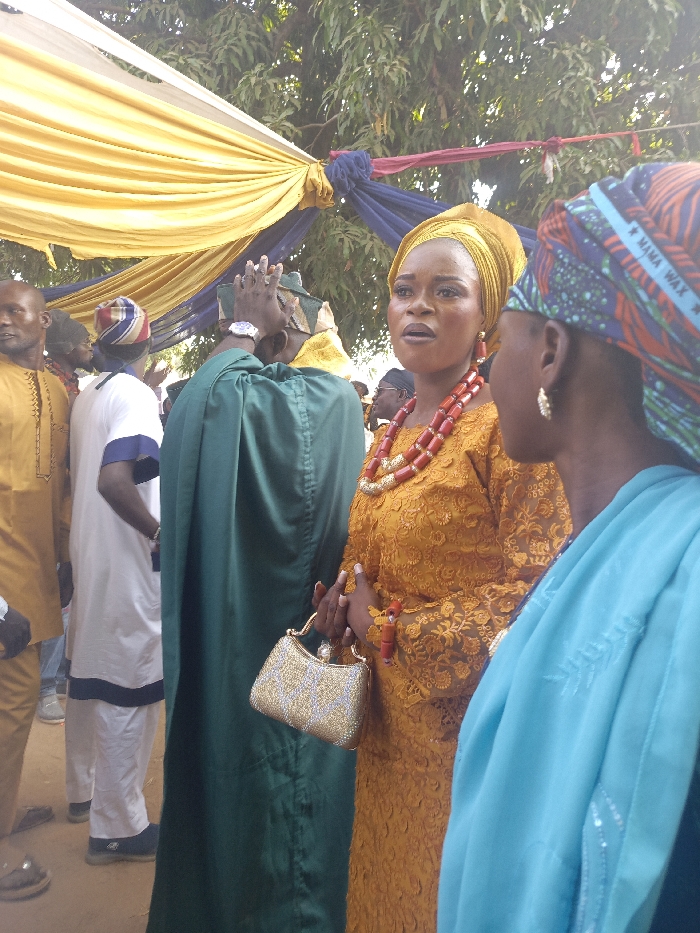 The image shows a group of people outdoors, possibly at a wedding or celebration. A woman in a yellow lace dress, head wrap, and red beaded necklace holds a silver purse and appears to be speaking. Another woman with a headscarf is beside her, partially obscuring the view. Behind them, people are dressed in traditional attire, including green robes and white outfits with blue trim. Yellow and blue fabrics hang overhead.