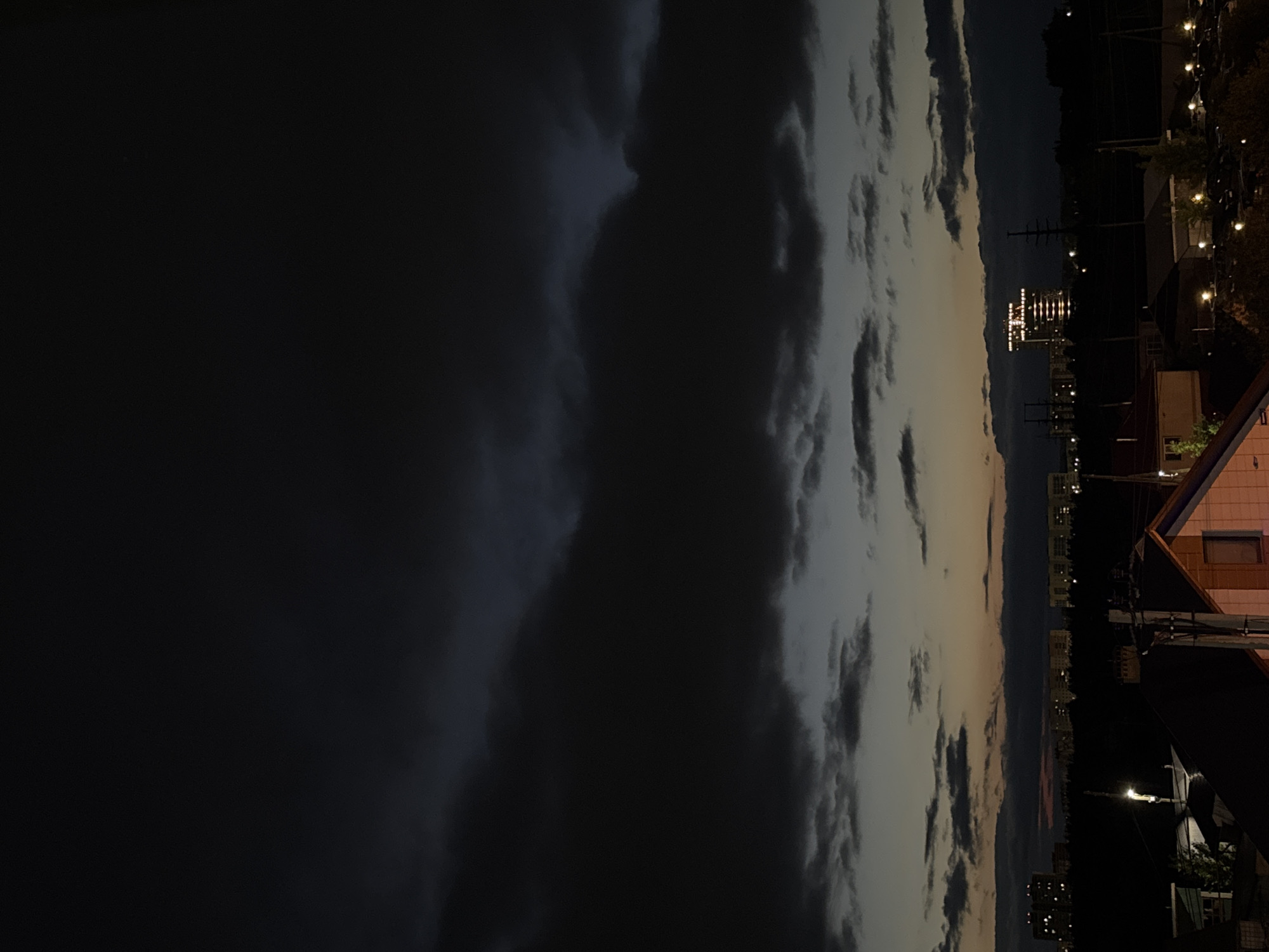 The image depicts a twilight scene over the city of Kondratovo, Russia. The sky is dominated by heavy, dark clouds, suggesting impending rain or a storm. Below the clouds, a gradient of light transitions from deep blue and gray in the upper sky to a faint orange and yellow glow near the horizon. A line of buildings, illuminated by lights, stretches across the middle ground, appearing as a silhouette against the dimly lit sky. Among these buildings, one prominent structure stands out with a series of bright lights outlining its upper floors, possibly indicating an office building or residential complex. In the foreground, the rooftops of several houses are visible, with some lights on within the structures. The overall atmosphere is one of a darkening evening in a suburban or semi-urban environment. No people or specific actions are discernible in the image.