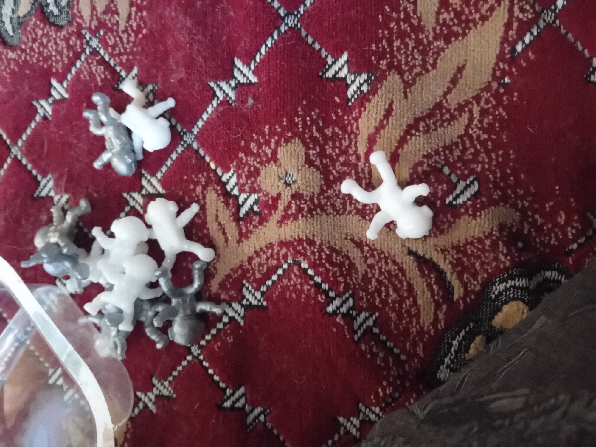 The image depicts a collection of small, plastic figurines, resembling babies, scattered across a red and tan patterned fabric surface. Some figurines are white, while others are gray. They are positioned in various orientations, suggesting they have been dropped or are being played with. In the upper left corner, a clear plastic container or bag is partially visible, also containing some of the figurines. The fabric appears to be a rug or blanket with a repeating geometric and floral pattern. There are no people clearly visible in the image, and the context of the location in Chuhuiv, Ukraine, is not visually determinable from the provided image. The scene is a still life arrangement of toys on a textured surface.