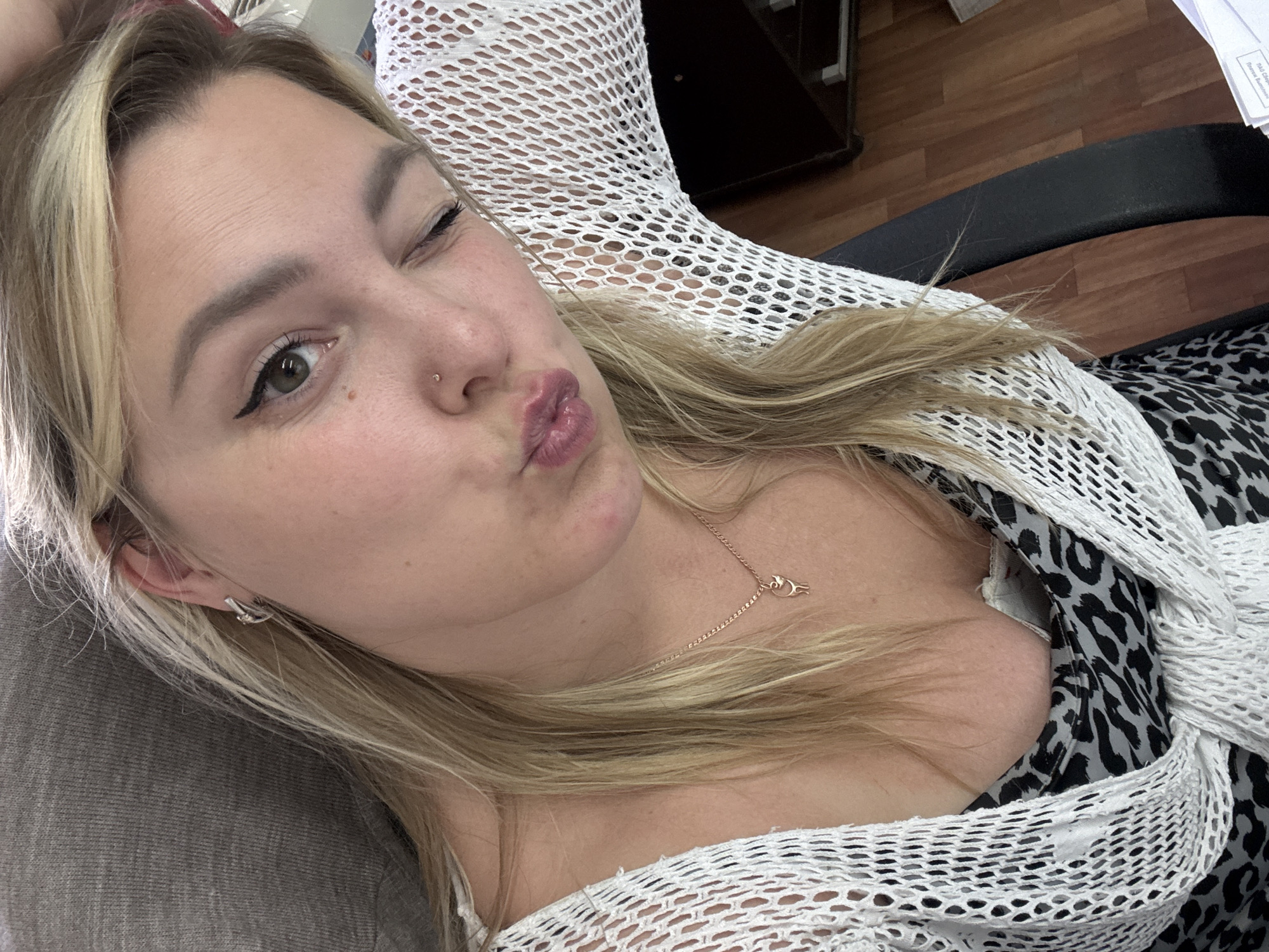 A young woman with blonde hair is taking a selfie in what appears to be an office setting. She is leaning back in a chair, with her left arm resting behind her head. She is wearing a white open-knit cardigan over a leopard-print top. The woman is winking and puckering her lips. Her hair is long and blonde, falling over her shoulders. She is wearing a small silver hoop earring in her left ear and a delicate gold necklace with a small pendant. She has a nose ring. The background includes a grey upholstered chair, a wooden floor, and a dark wooden desk. The lighting suggests an indoor environment, possibly during daylight hours. There are no discernible landmarks to place this image specifically in the city of Noviy, Russia.