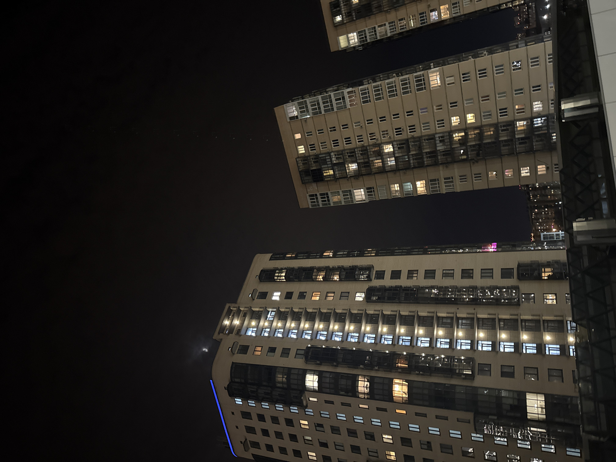 The image presents a striking, low-angle view of modern high-rise buildings against a dark night sky in Krasnoyarsk, Russia. The perspective looks sharply upwards, emphasizing the towering scale of the architecture. Several multi-story structures, likely apartment blocks or offices, dominate the frame, their facades characterized by numerous windows. Many of these windows are brightly illuminated, casting warm and cool light onto the dark surrounds and suggesting active occupancy within the buildings.
The architecture is contemporary, featuring repetitive grid patterns of windows and some external elements such as balconies or decorative structures. On the left side of the frame, a faint crescent moon is visible in the clear, black sky, indicating nighttime and fair weather. A distinct bright blue light strip runs vertically along the bottom-left edge of one of the buildings, adding a modern, perhaps decorative, touch to the urban landscape.
No specific activities or events involving people are explicitly shown, but the glowing windows convey the ongoing life within the city after dark. The composition highlights the verticality and density of the urban environment at night. There is no visible text in the image.