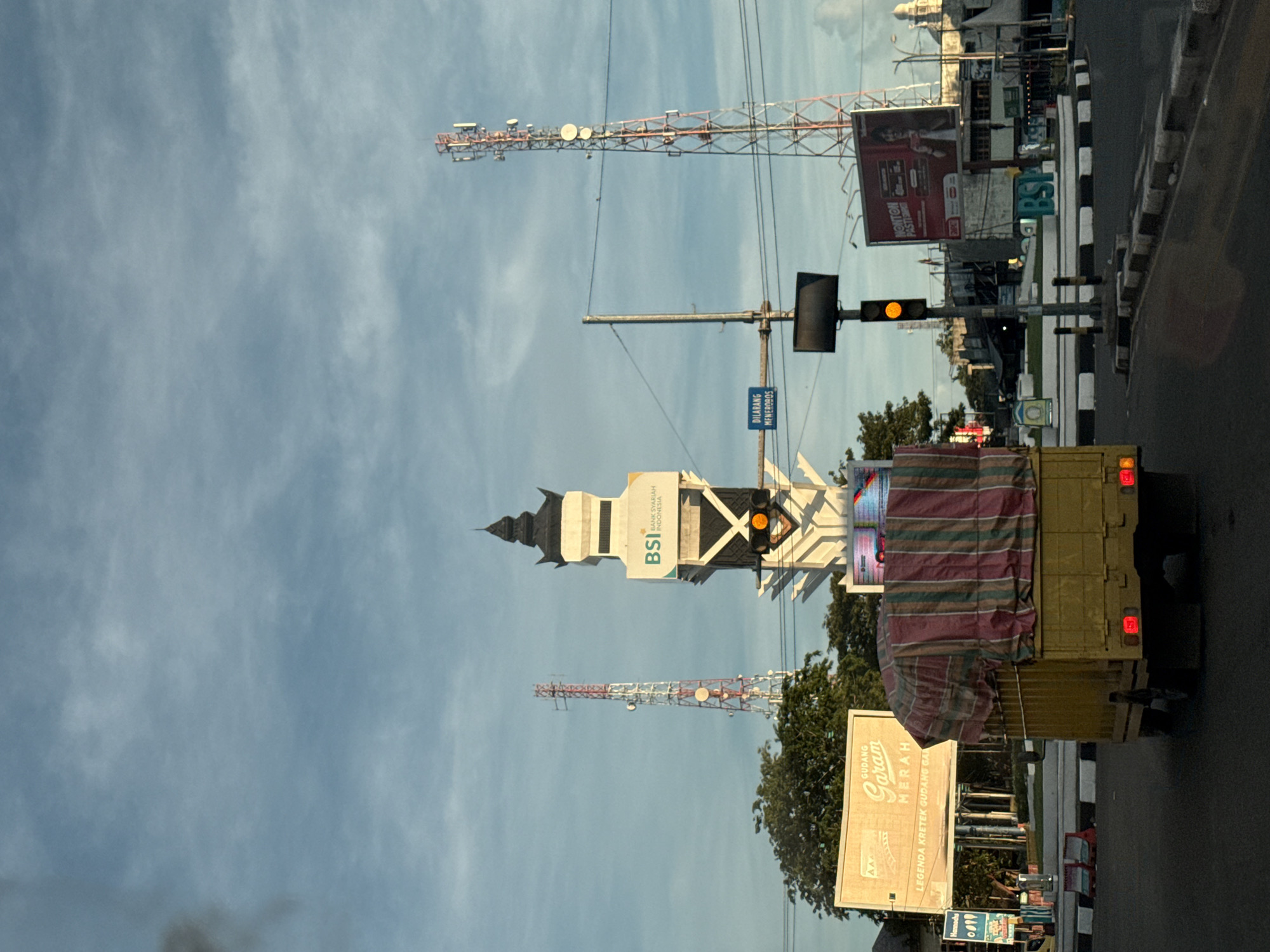 The image shows a street scene in Lambaro, Indonesia, with a yellow truck partially obscuring the view. A tall BSI (Bank Syariah Indonesia) building is visible, along with telecommunication towers in the background. The sky is cloudy. Street signs and billboards for brands like "Gudang Garam Merah" are present. A traffic light shows a yellow light. There is a blue sign that says "DILARANG MENEROBOS"
---