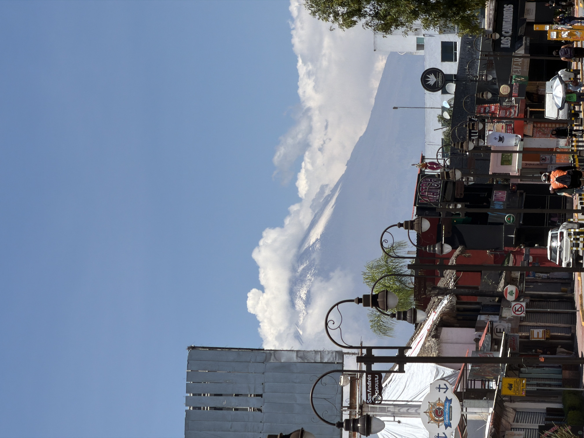 The image depicts a street scene in San Andrés Cholula, Mexico, with a prominent mountain visible in the background, partially obscured by clouds. The foreground features various street elements, including street lamps and signs. Numerous commercial signs advertise local establishments. People are present on the street, though they are not the primary focus. The sky is clear and blue.