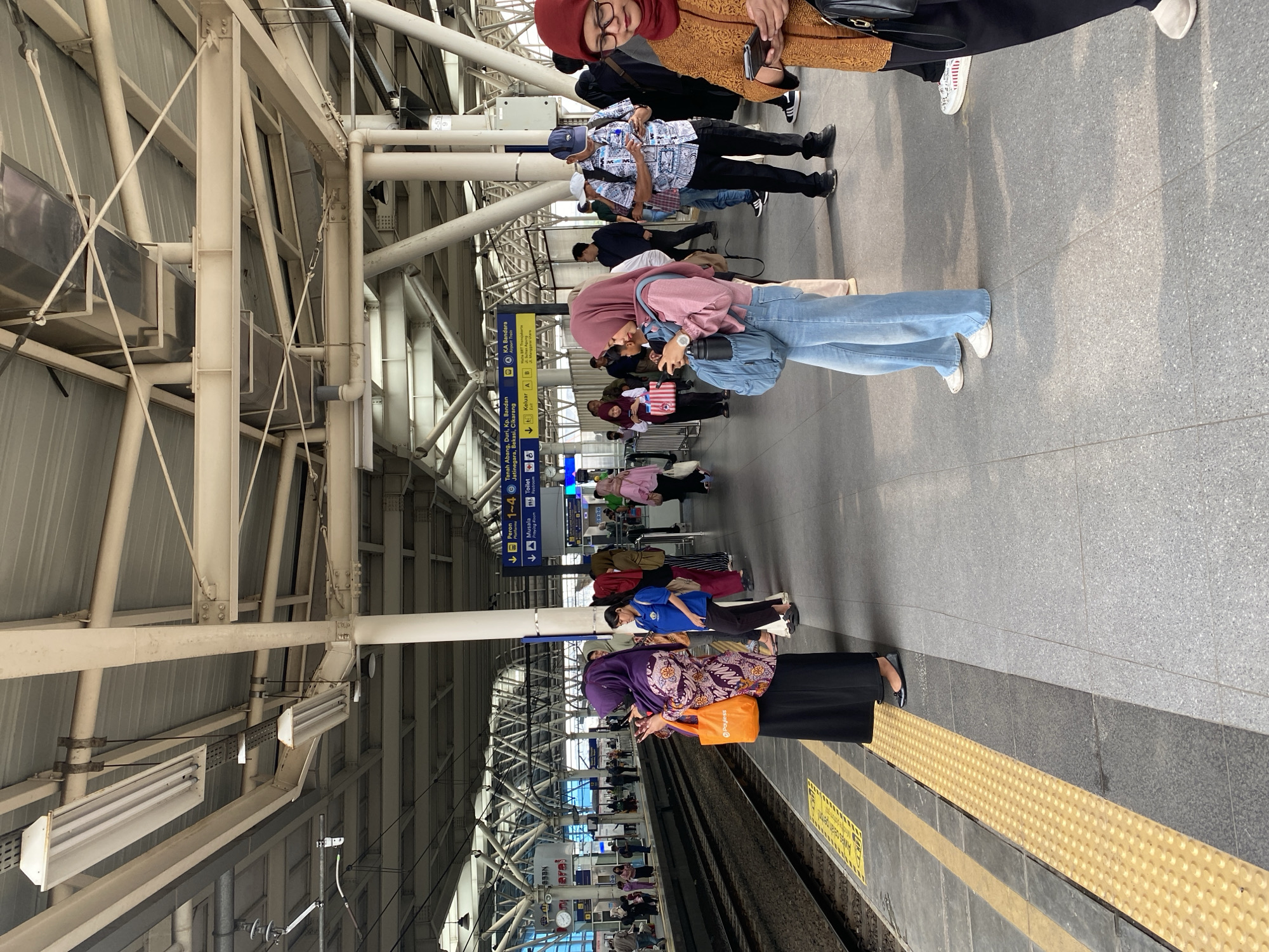 The image depicts a busy train platform at Manggarai Station in South Jakarta, Indonesia. Several individuals, predominantly women wearing hijabs, are standing on the platform, many of them looking at their mobile phones. One woman in the foreground, wearing a purple hijab and a patterned top, holds an orange bag. Beside her, another woman in a pink hijab and light blue jeans is also engrossed in her phone, with a backpack at her feet. A man in a patterned shirt and a blue cap stands further down the platform, also looking at his phone. The platform itself is paved with grey tiles, with a textured yellow safety strip running along the edge closest to the train tracks. Overhead, a complex network of steel beams and light fixtures supports the station's roof. Signs indicating platform numbers and destinations are visible in the background. The scene appears to be a typical moment of transit, with people waiting for their trains.