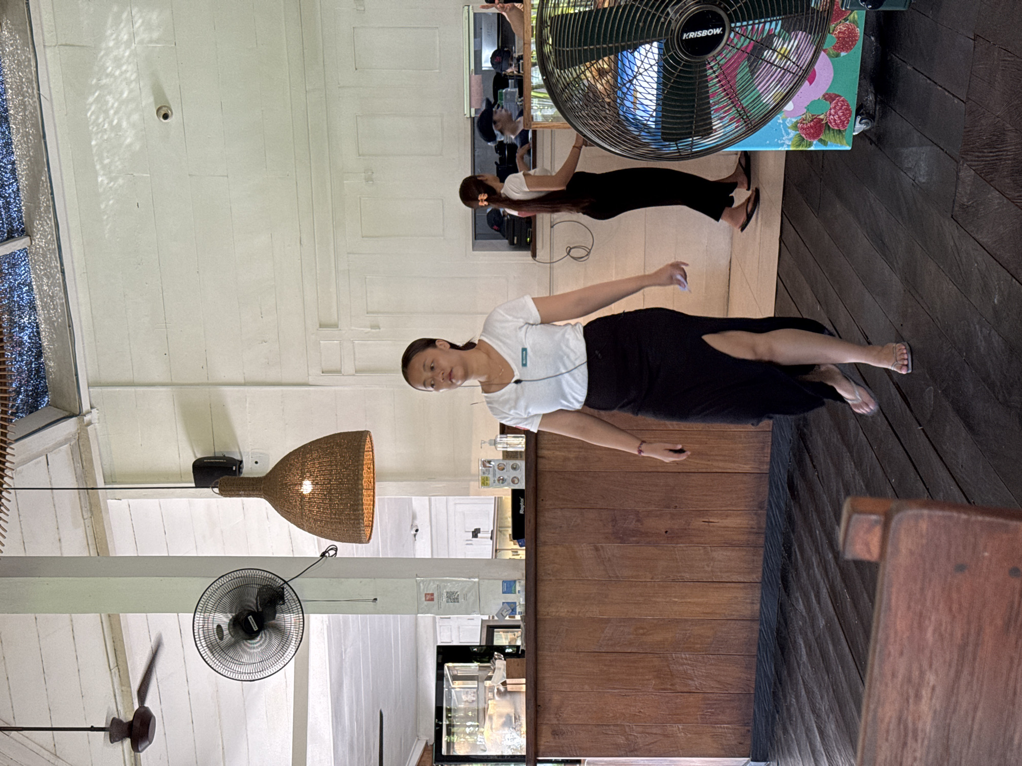 This image captures an indoor or semi-outdoor space, likely a cafe or restaurant, characterized by a rustic and airy aesthetic, consistent with its North Kuta, Indonesia location. The setting features light-colored paneled walls and ceiling, contrasting with a dark wooden floor and a substantial wooden counter or bar area. Natural light streams in, creating dappled patterns on the ceiling.
Two women, possibly staff members, are visible. The woman in the foreground stands facing slightly towards the camera, wearing a white short-sleeved top, a long dark skirt with a side slit, and light-colored flip-flops. She has dark hair pulled back and a neutral expression, with a white earphone wire visible on her chest. A small, unidentifiable white badge is pinned to her shirt. The second woman is further back, behind a counter, wearing a white top and dark trousers. Her reflection in a mirror shows her wearing a cap.
Various objects are integrated into the space. Multiple fans are present, suggesting a warm climate: a large, wall-mounted "KRISBOW" brand oscillating fan with a colorful base featuring raspberry graphics, a smaller wall-mounted fan, and a ceiling fan with wooden blades. A large, basket-weave pendant light fixture hangs prominently. Other details include a small security camera and a speaker on the ceiling, bottles (possibly hand sanitizer) and other items on the counter, and a display case behind the counter. A section of shimmering, reflective material is visible near a window on the left. The overall atmosphere appears calm and functional.