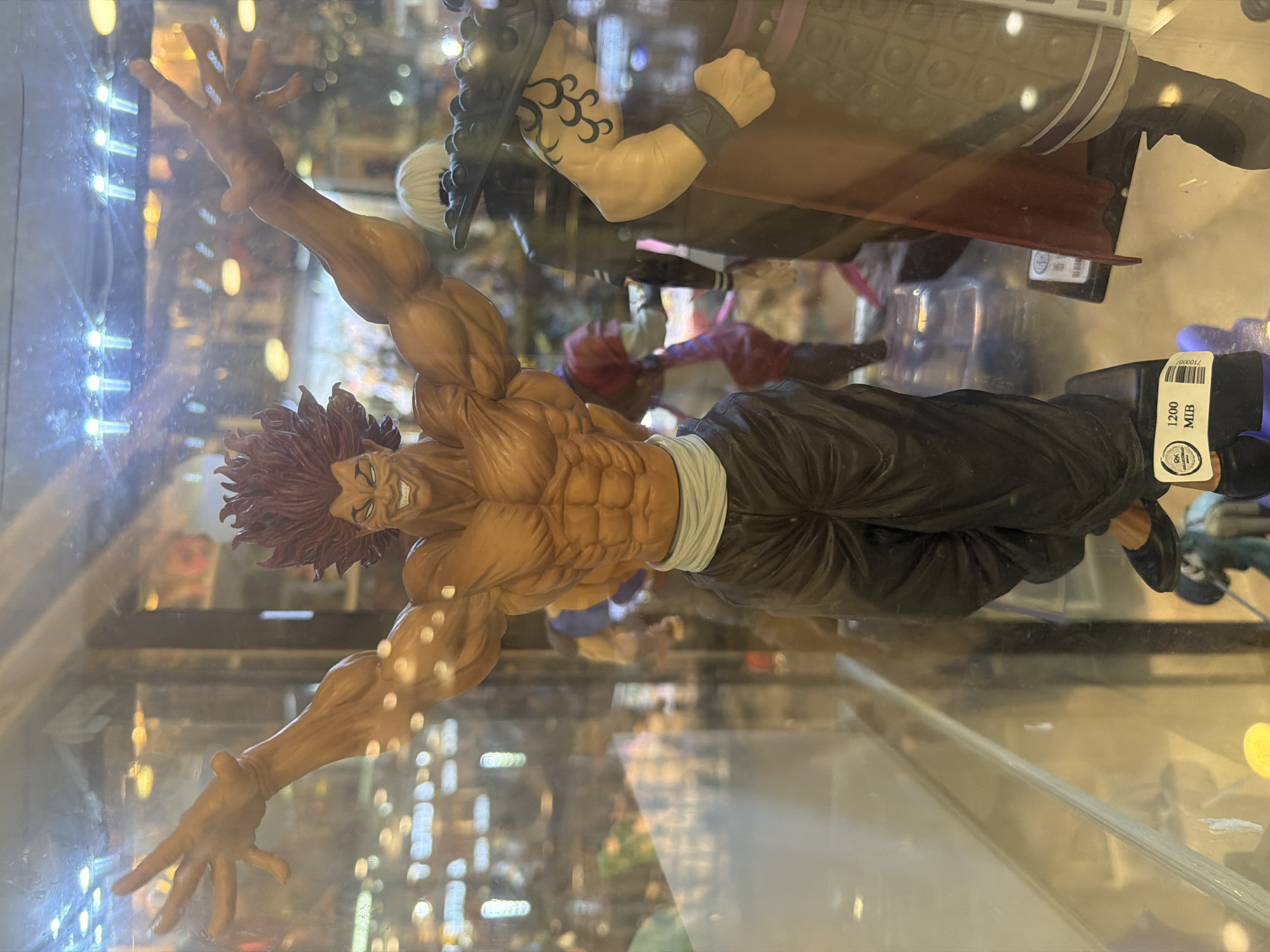 A well-lit indoor display case contains several anime figurines. The primary focus is a large, muscular male figurine with spiky red hair, a wide grin, and arms outstretched. He is wearing dark pants and a white waistband. To the right, partially obscured, is another figurine with white hair and a dark outfit, and further right is a figurine with a brown garment and a tattoo on its arm. In the background, other figures and shelves with merchandise are visible. The display case appears to be in a retail environment, possibly a collectibles shop. A price tag is visible on the base of the main figurine, reading "1200 MIB." Based on the provided information, this scene is located at RK Collectibles in Davao City, Philippines.